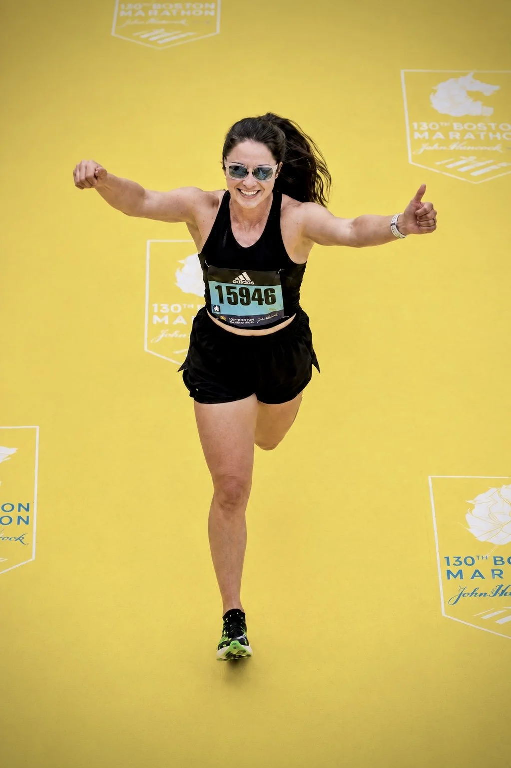 A woman running and smiling with her arms raised, participating in the 130th Boston Marathon, wearing sunglasses, a black athletic outfit, and a race bib number 15946, against a yellow background featuring marathon branding.