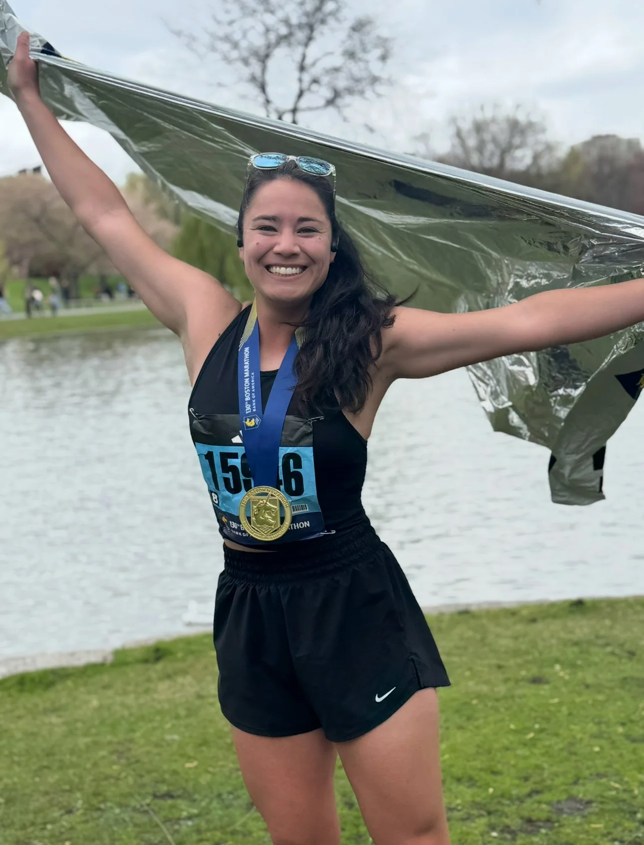 A smiling young woman in athletic clothing celebrating after completing a race, holding a reflective blanket and wearing a medal, with a pond and trees in the background.