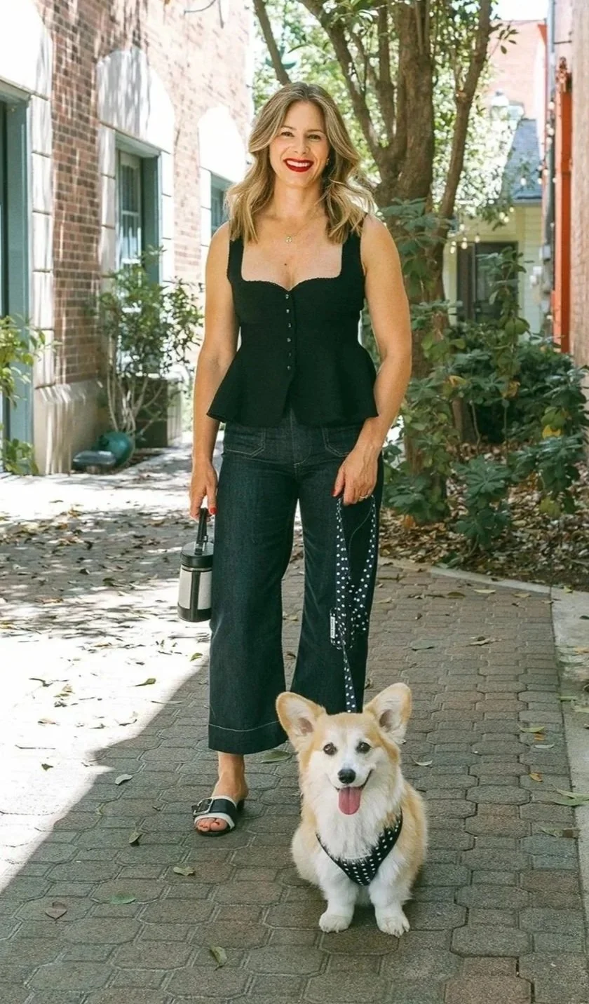 A woman with shoulder-length blonde hair, smiling and wearing a black sleeveless top, dark wide-leg jeans, and black sandals with white accents, is standing outdoors on a sidewalk with a corgi dog that has a colorful coat and a polka dot harness. The woman holds a black water bottle, and the dog has its tongue out, looking happy.