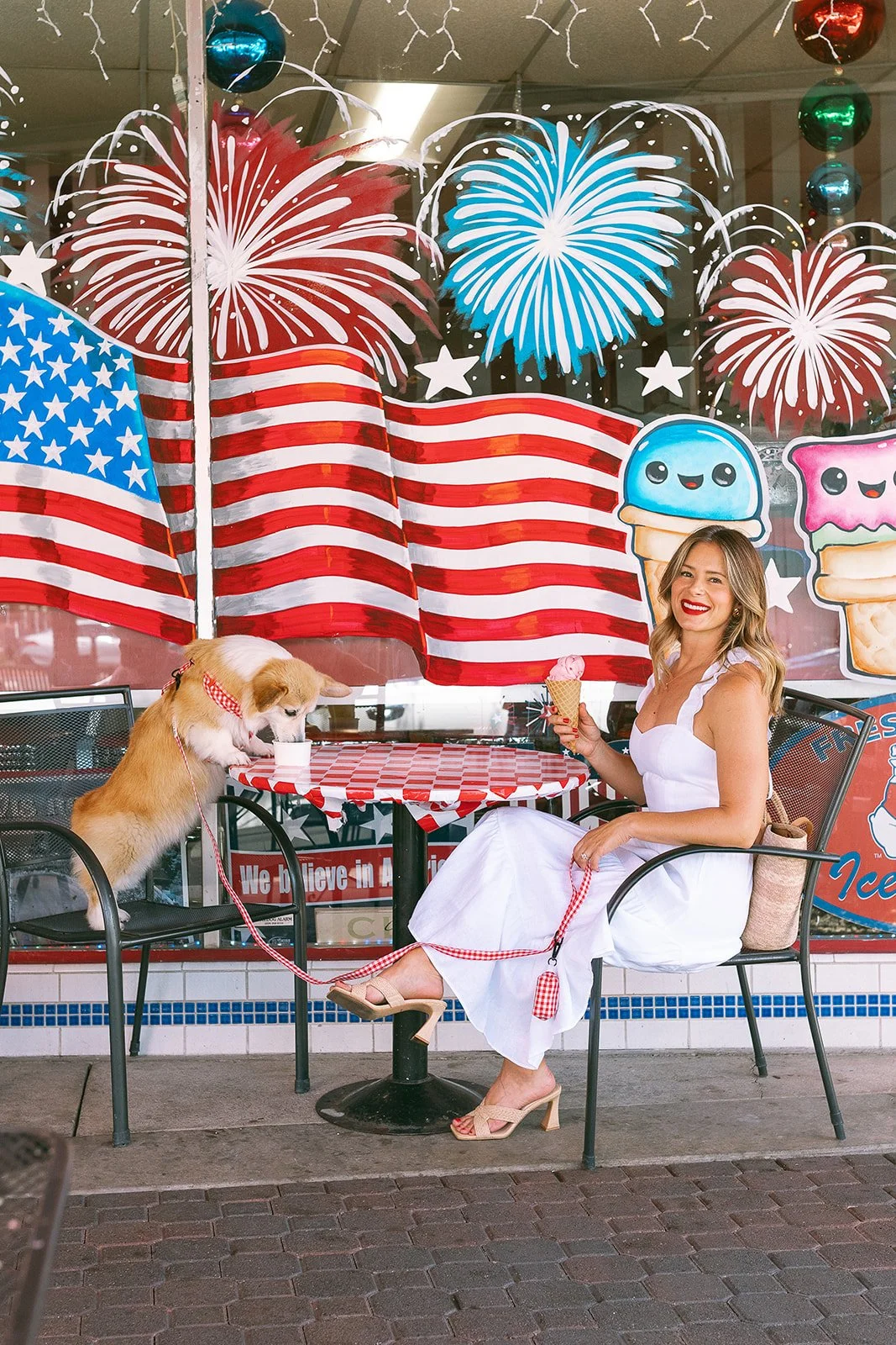 A woman sitting at an outdoor table with a dog, holding an ice cream cone, in front of a window decorated with American flag, fireworks, stars, and cartoon ice cream graphics for Independence Day celebration