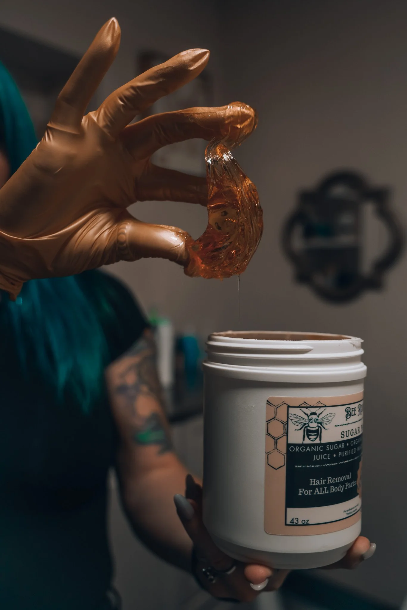 A person wearing gloves is holding a large container of organic sugar, which is labeled for hair removal for all body parts. Sugaring ball