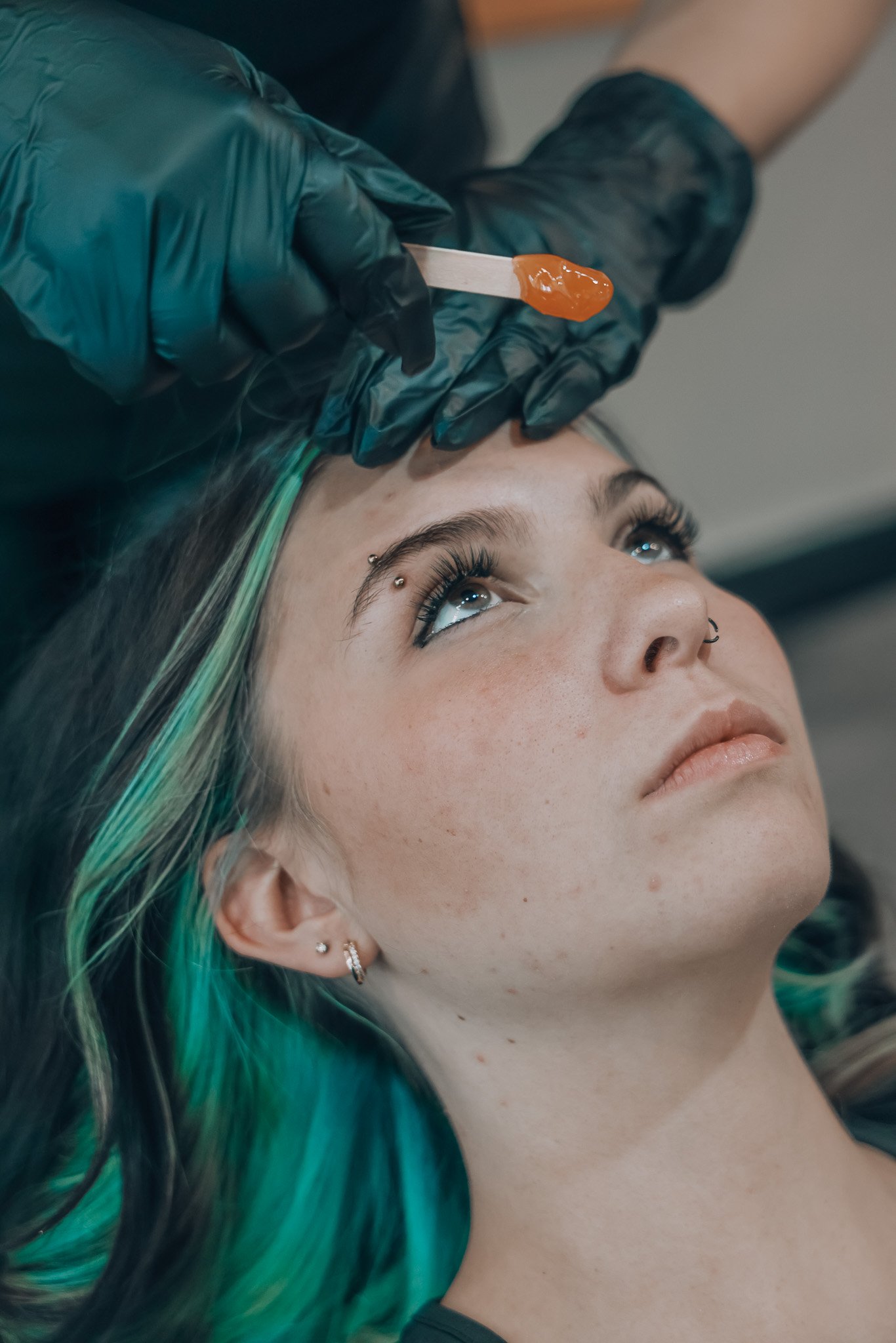 A person with green and black hair lying down while a tattoo waxer in gloves applies wax to eyebrows for an eyebrow wax