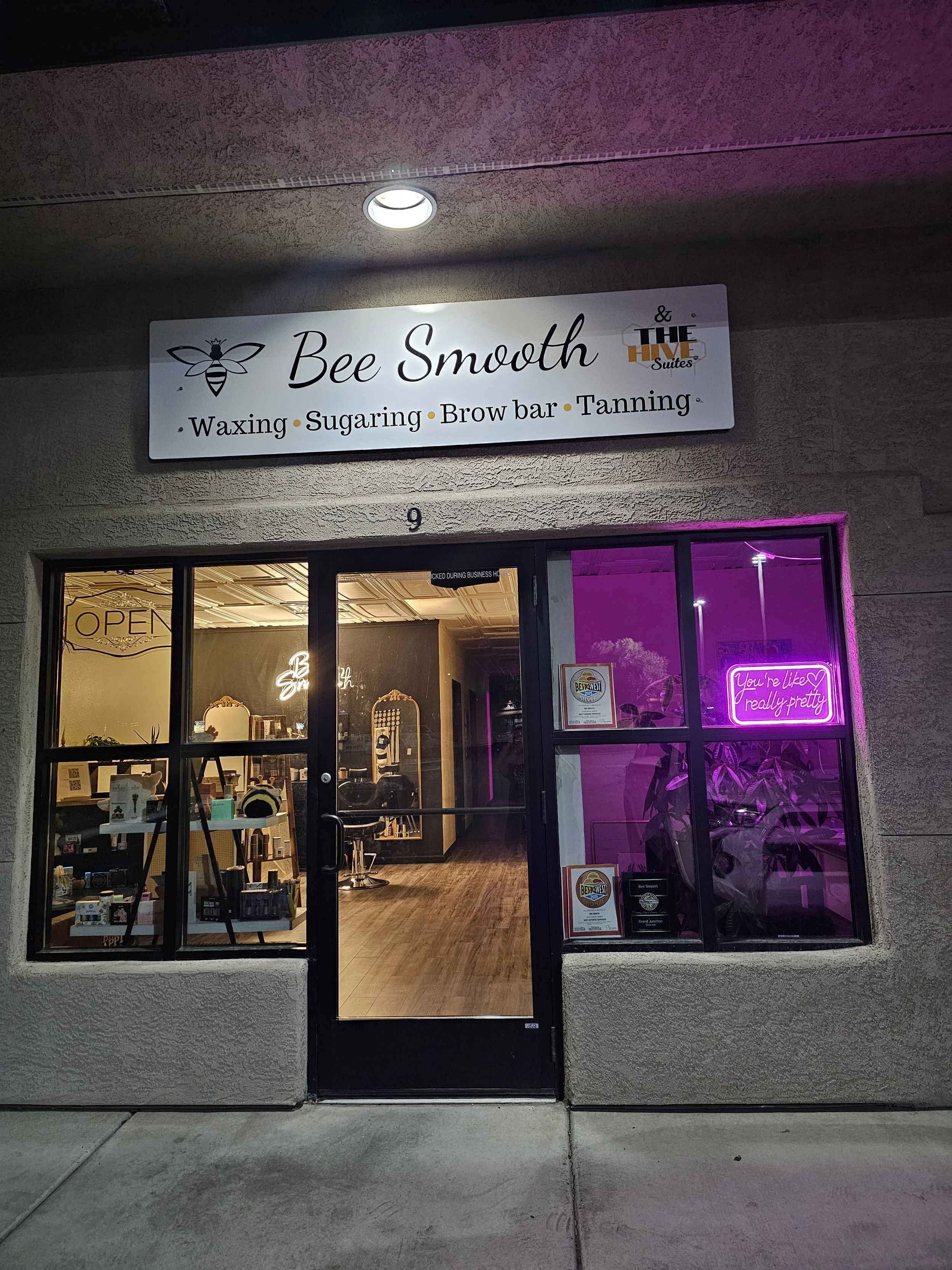 The storefront of Bee Smooth, a beauty salon offering waxing, sugaring, brow bar, and tanning services. The store has a sign with a bee illustration and the name 'Bee Smooth' in cursive lettering. The storefront includes a window displaying the interior, a neon sign that reads 'You're like really pretty,' and various decorations.