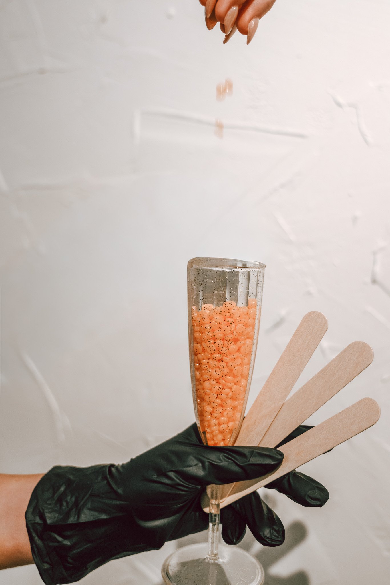 Person holding a tall glass filled with orange wax beads, using a black glove, against a white background. Several wooden popsicle sticks are fanned out in front of the glass. Waxing picture