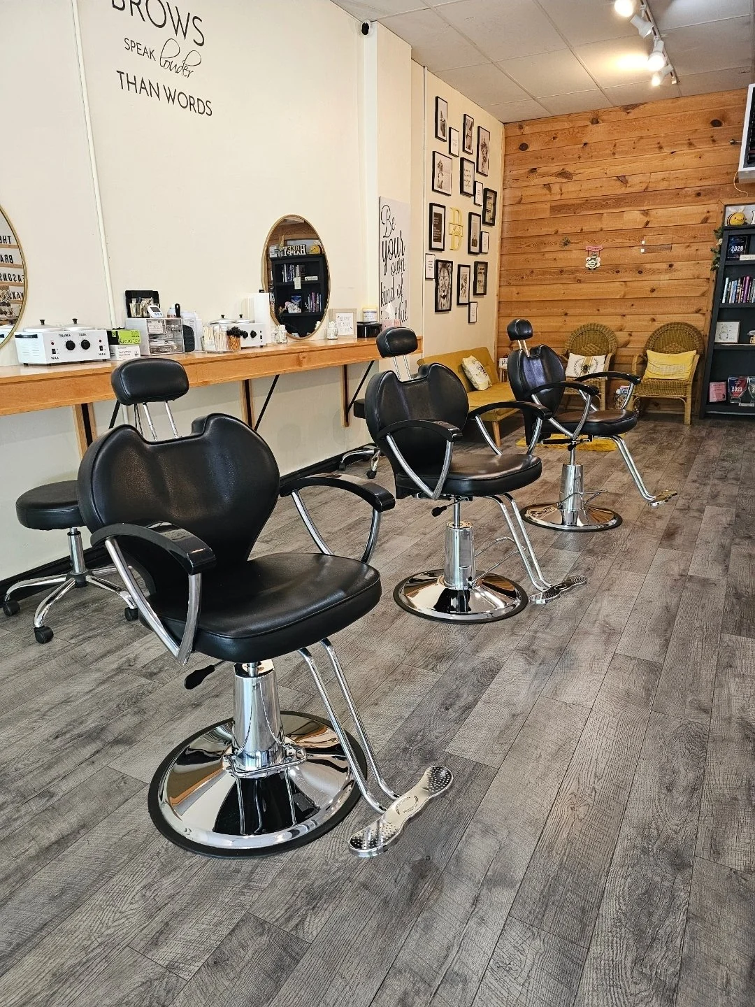 Waxing salon with three black salon chairs facing a long wooden counter with mirrors and waxing products. The wall has framed art and motivational quotes, and there are wicker chairs and a bookshelf in the background. inside Bee Smooth Main street