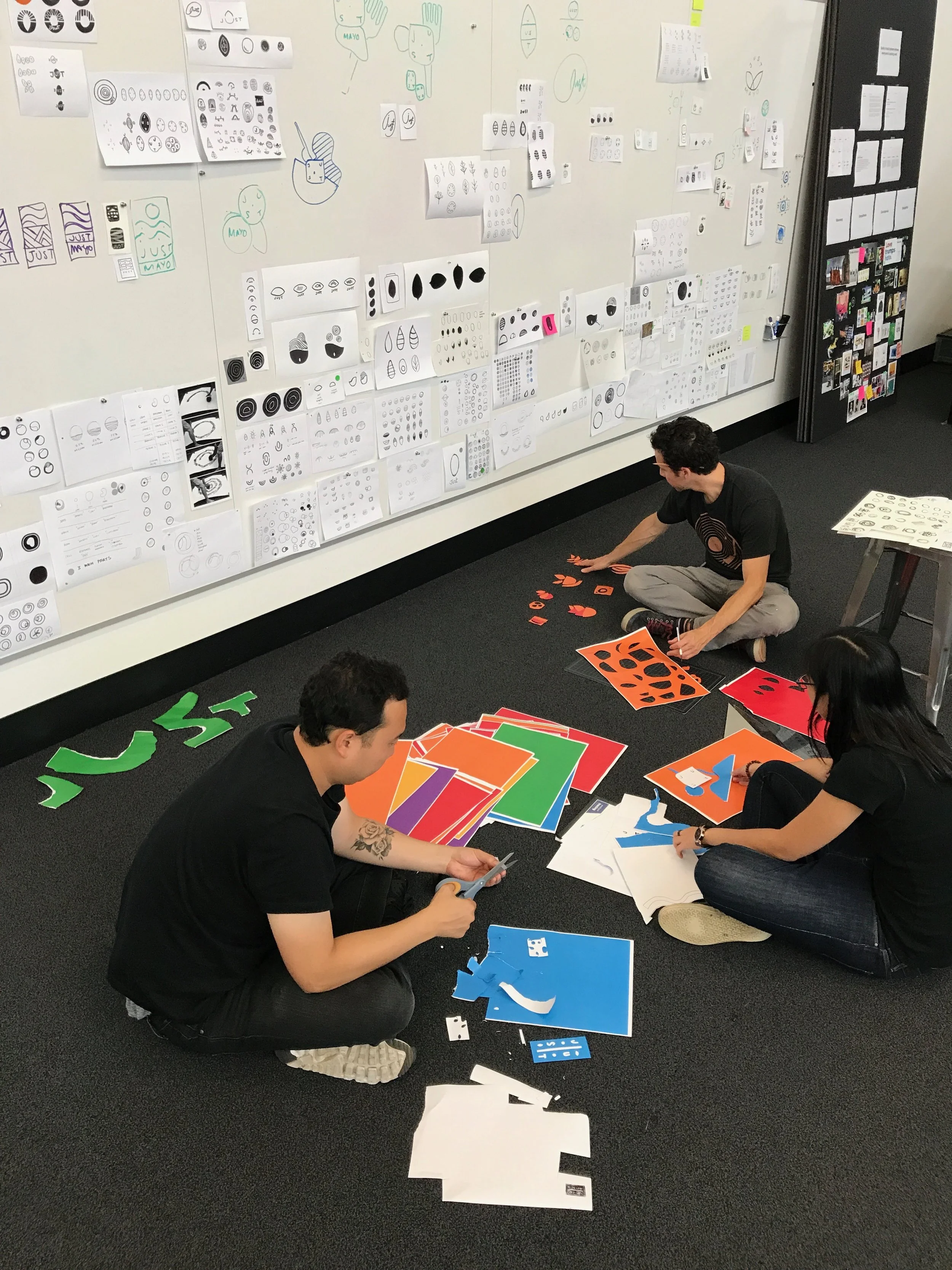 People sitting on the floor working on colorful paper cut-outs with various shapes and patterns, in a room with whiteboard walls covered in sketches and designs.