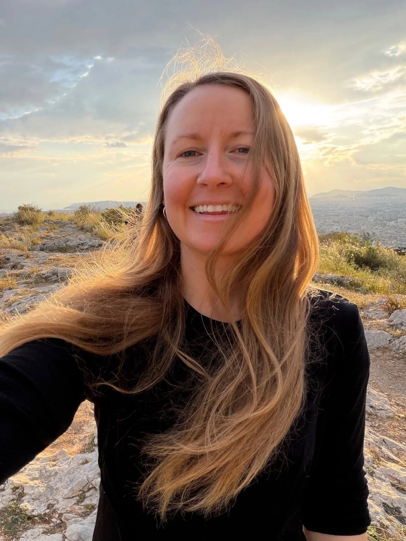 A woman with long wavy light brown hair smiling outdoors at sunset, wearing a black top, with rocky terrain and a distant cityscape in the background.