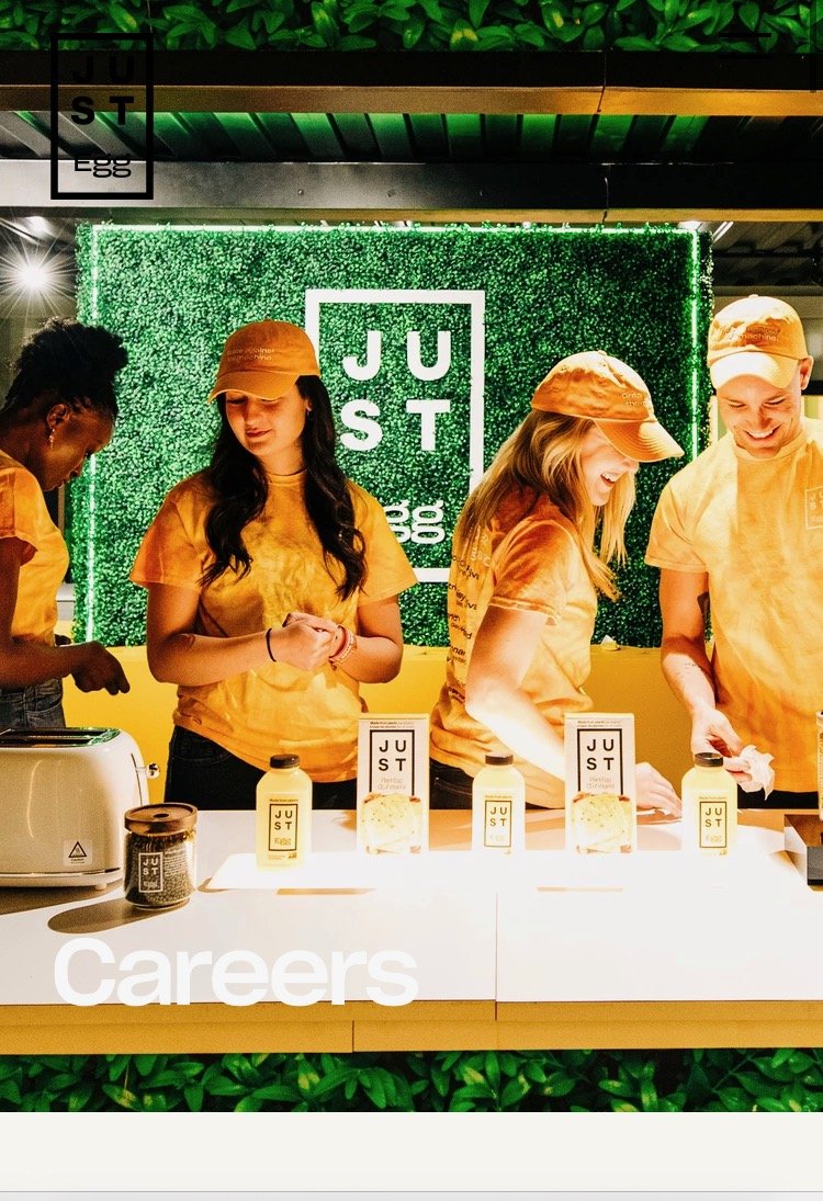 Four staff members wearing matching yellow shirts and hats working behind a counter at an event, with a green textured backdrop and a sign that says "JUST" in the background.