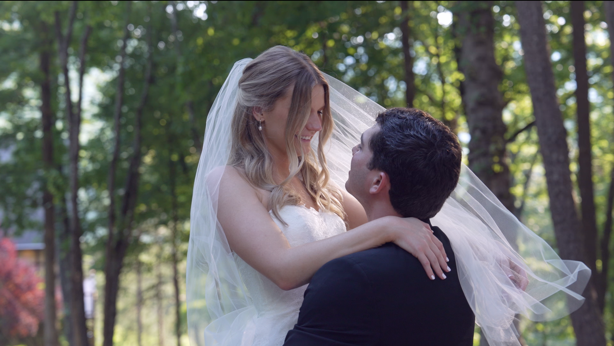 A bride and groom embracing outdoors in a wooded area during daytime. The bride wears a white wedding dress and veil, and the groom is in a black suit. The bride is smiling and looking at the groom, who is holding her up.