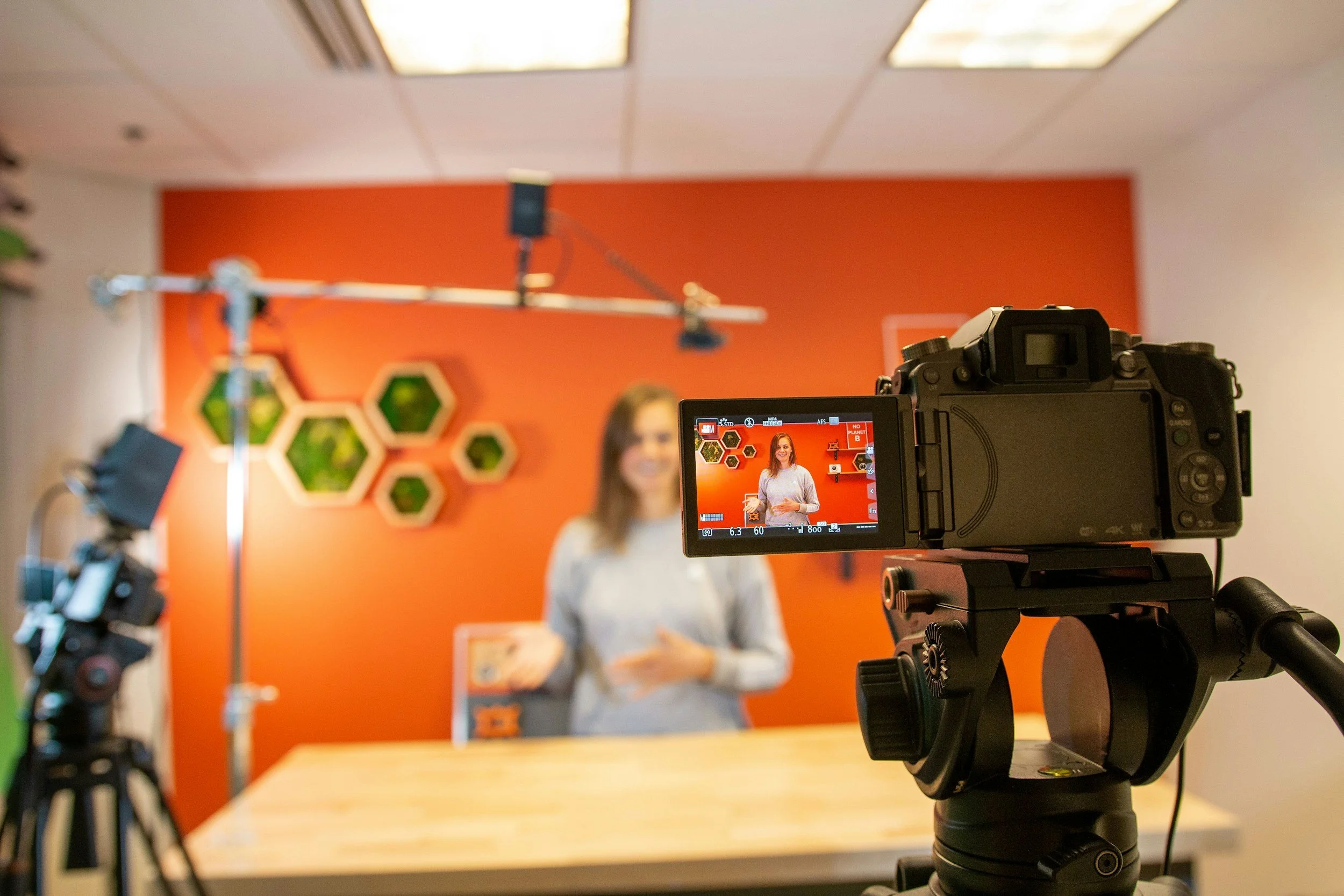 A professional video camera on a tripod recording a woman in a studio with an orange wall and hexagonal decor in the background.