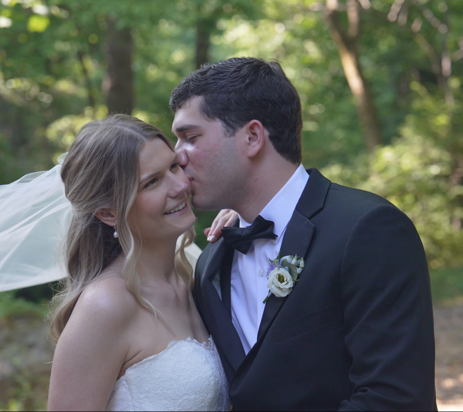 A bride and groom sharing a kiss outdoors in a wooded area during a wedding ceremony, with the bride in a white lace dress and the groom in a black tuxedo.
