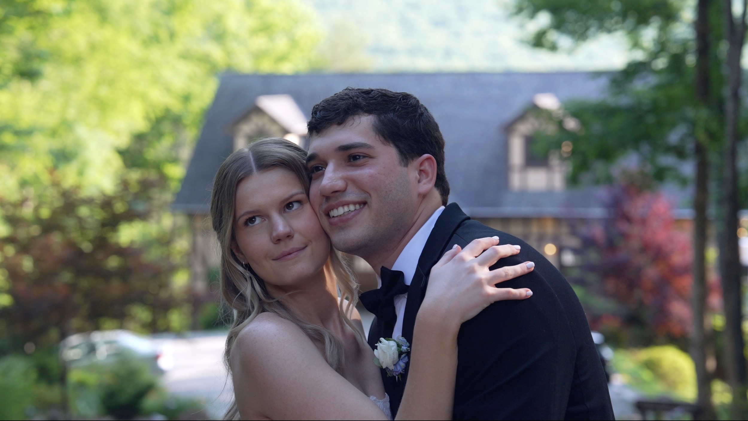 A couple dressed in wedding attire, embracing outdoors with a house and trees in the background.