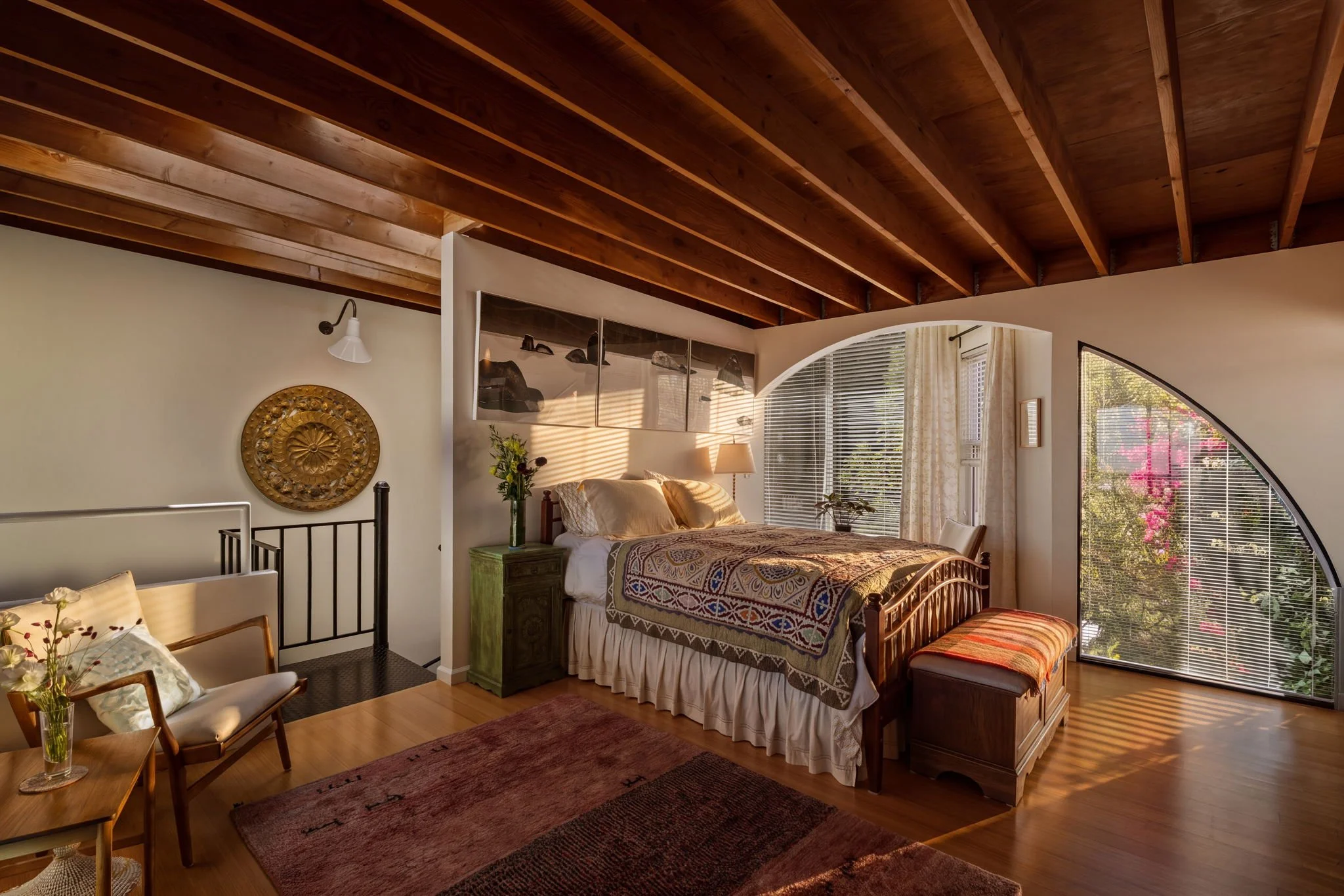 A cozy bedroom with a wooden vaulted ceiling, large arched window with blinds, and a bed with a colorful patterned bedspread. There is a nightstand with a lamp, a green dresser, and a wall decoration above the bed. The room has natural light streamin