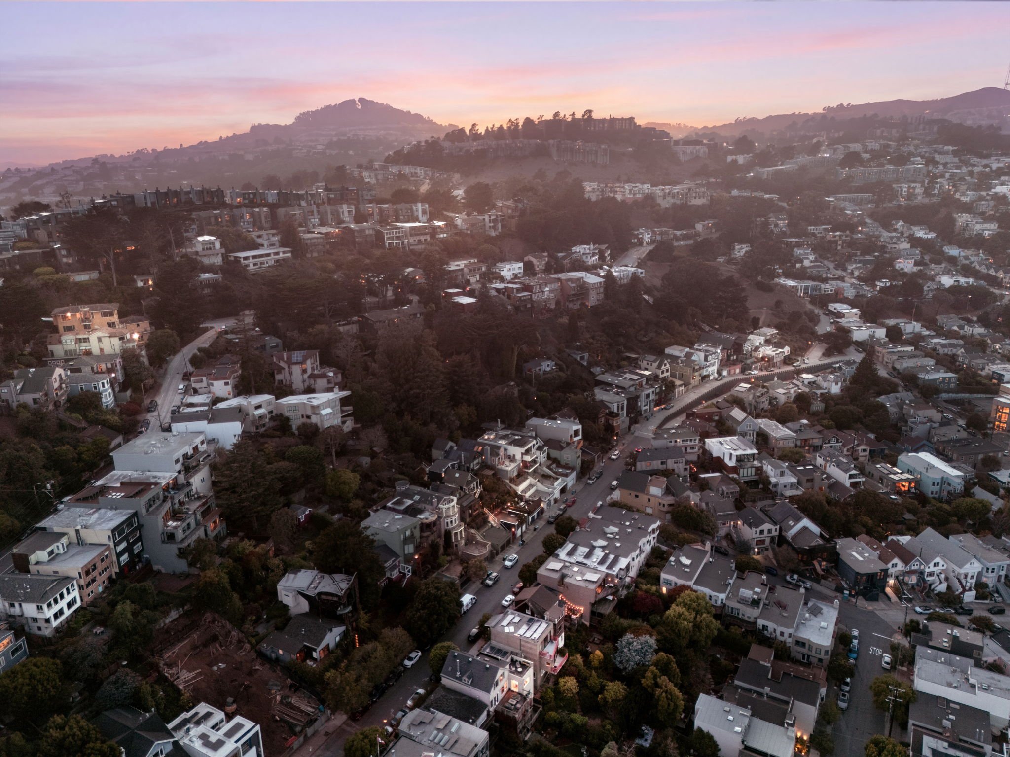 A hilly residential neighborhood at dusk with houses, trees, and a colorful sky.