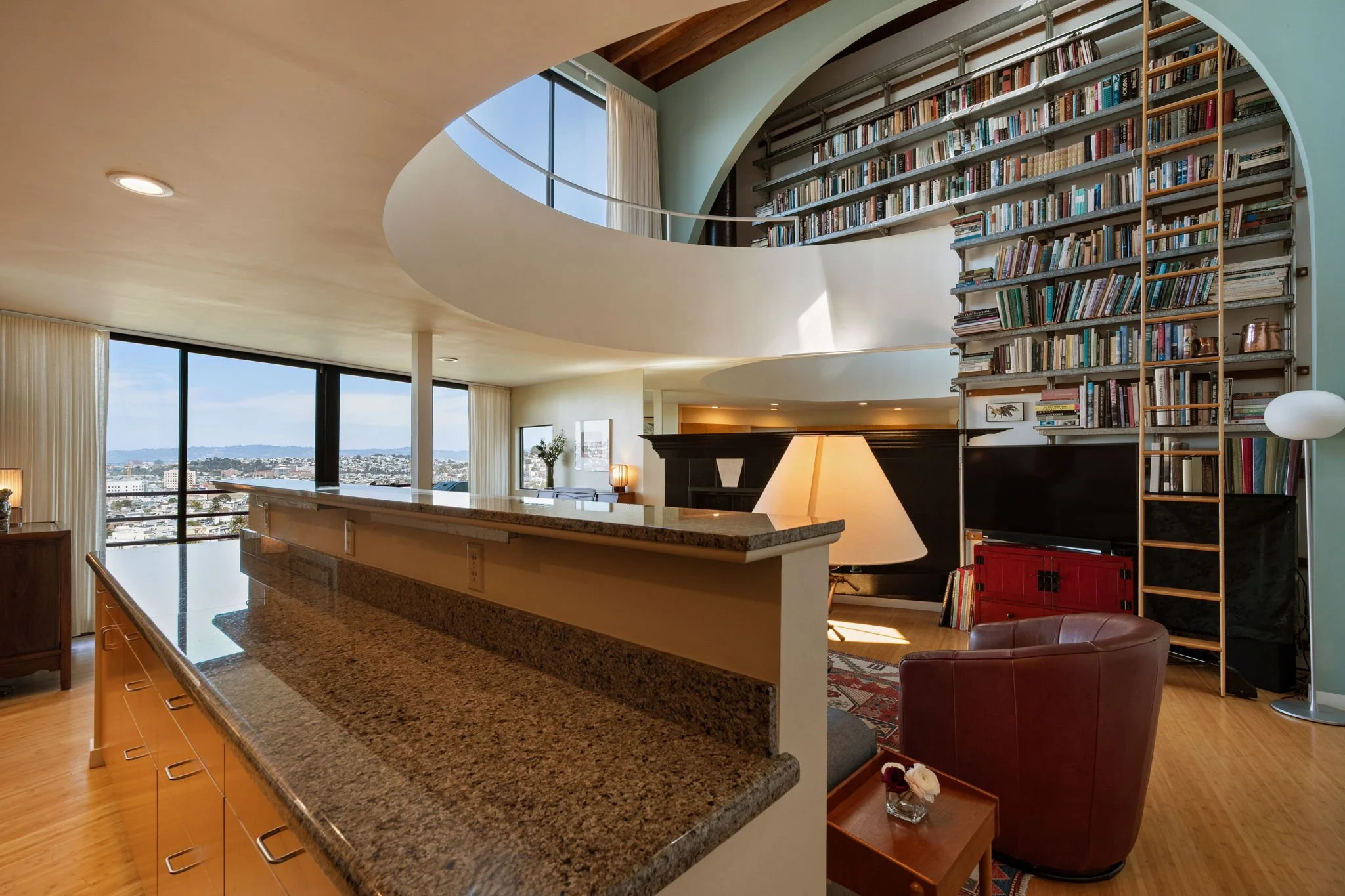 Interior view of a modern apartment with a curved bookshelf filled with books, large windows with curtains, and a kitchen counter with a granite top.