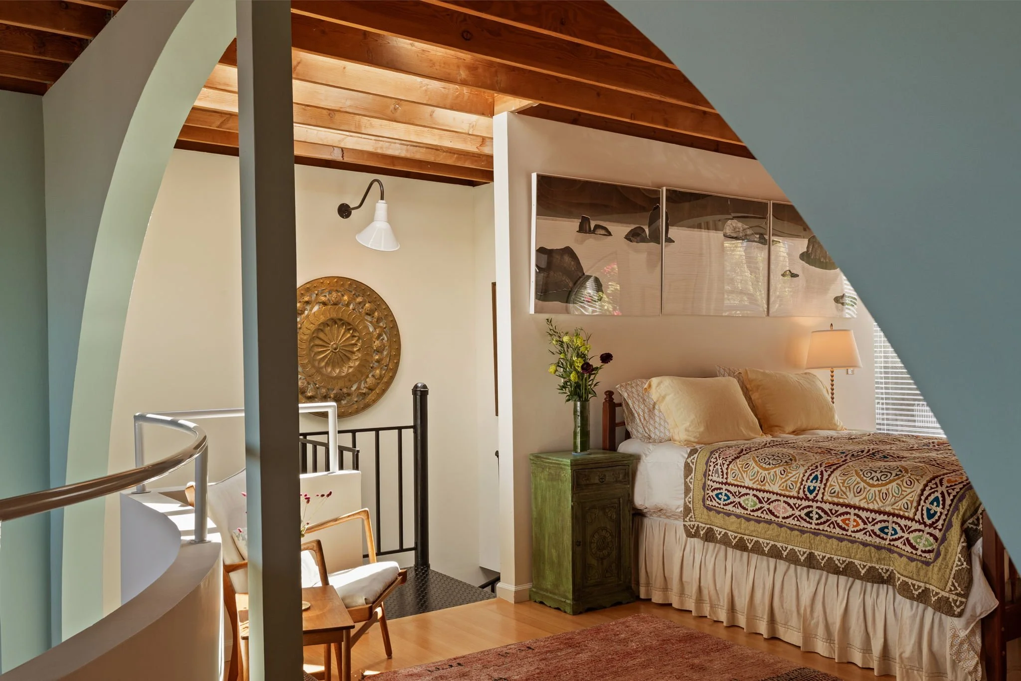 A bedroom with a vaulted wooden ceiling, a bed with a quilt, a nightstand with a vase of flowers, and a window with blinds. The photo is taken from behind a staircase railing.