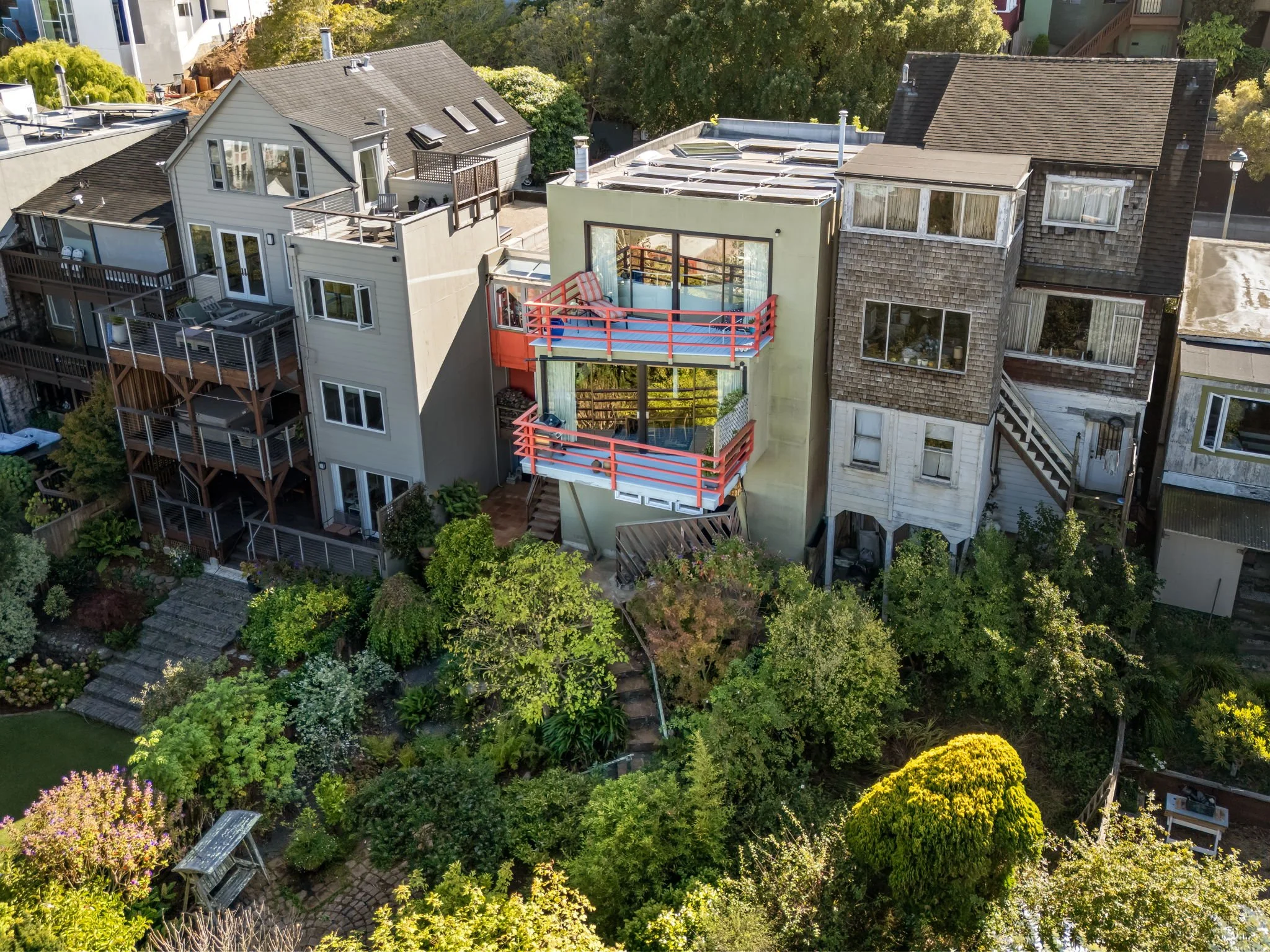 Aerial view of a neighborhood with multiple houses, including one with a unique structural design and blue and red balconies, surrounded by lush green trees and landscaped gardens.