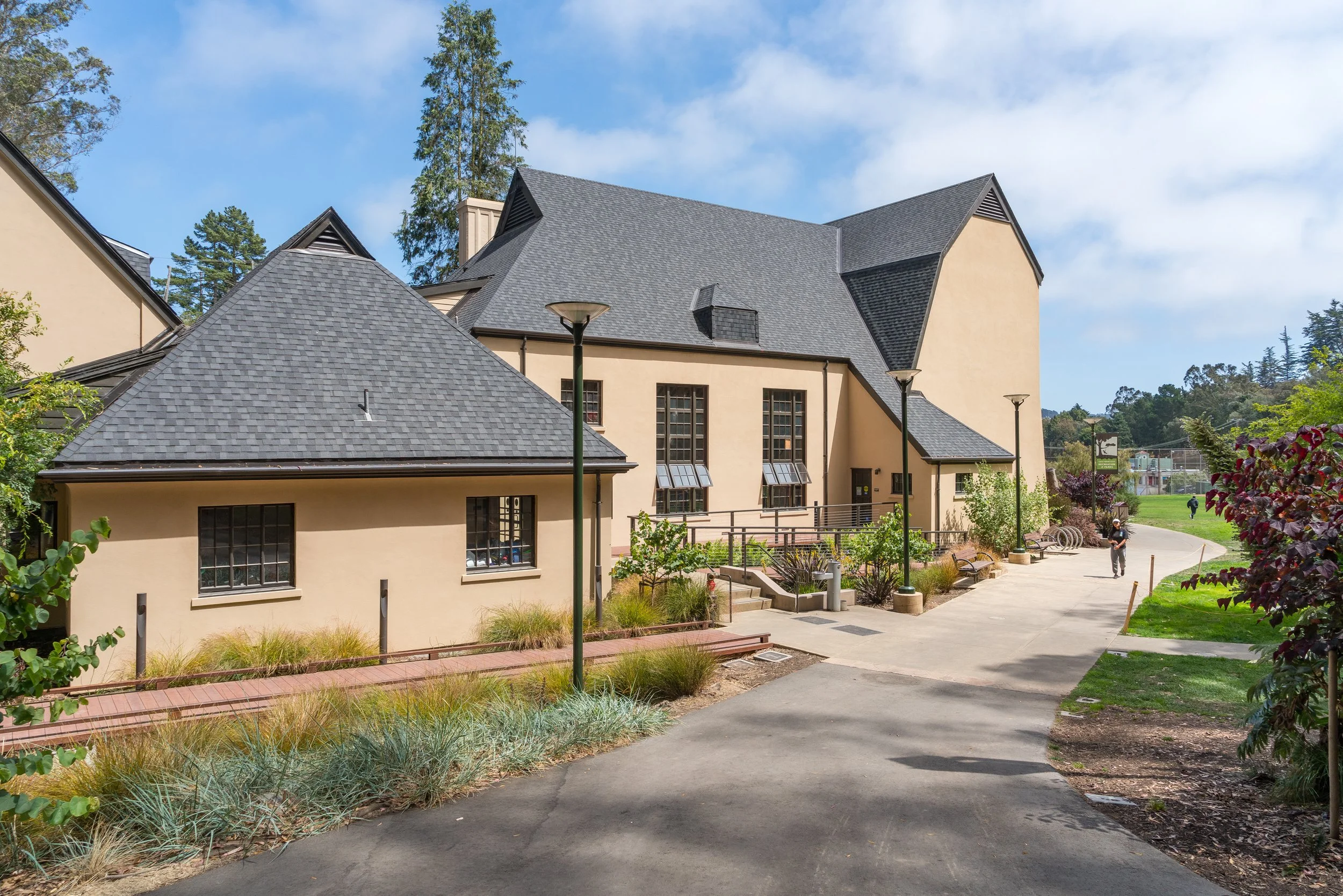 A pathway in a park leading past a cream-colored building with multiple roofs and windows, surrounded by green plants, trees, and street lamps under a partly cloudy sky.