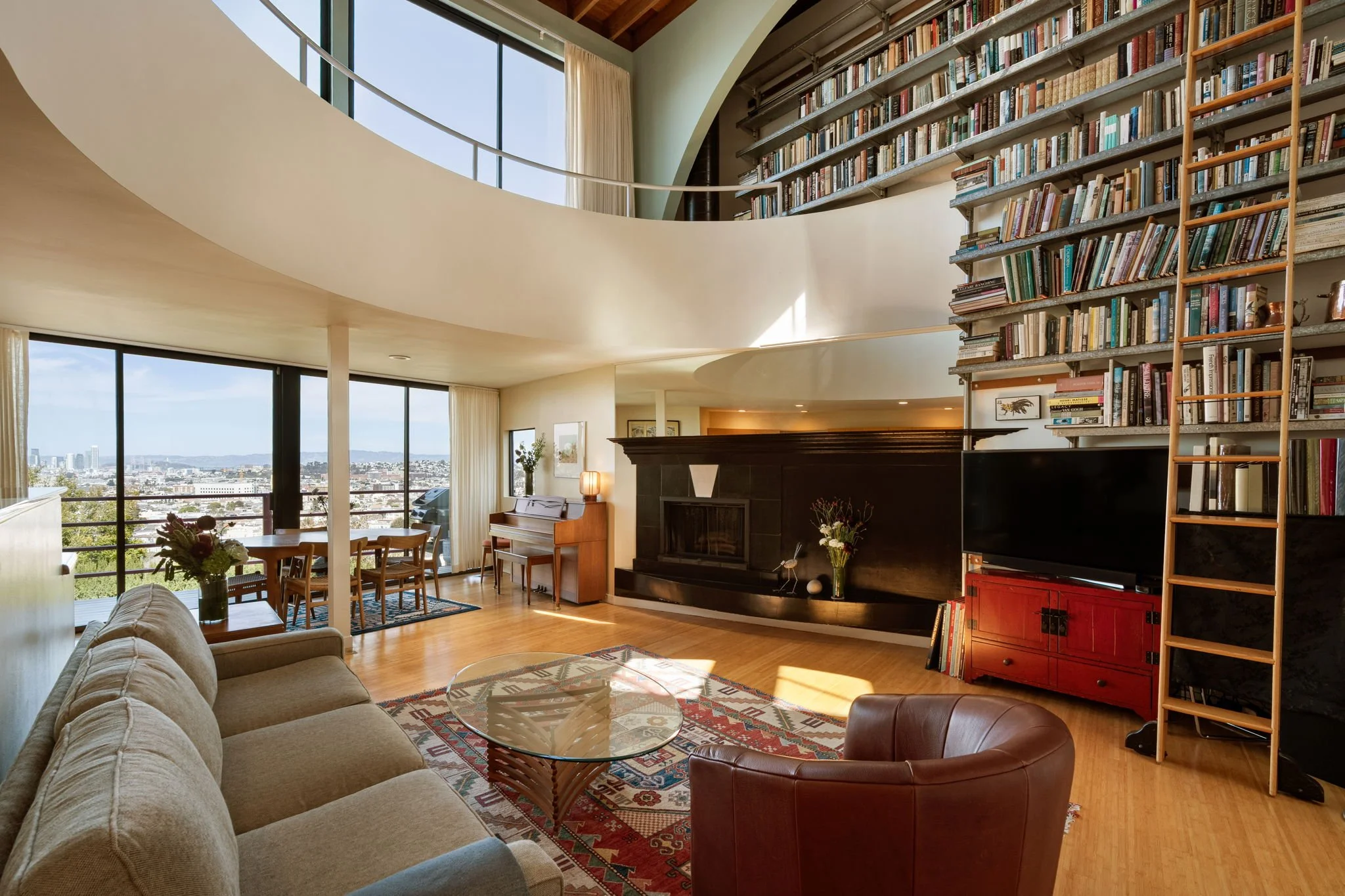 Interior of a living room with a large bookshelf, fireplace, piano, and a view of a cityscape through large windows.