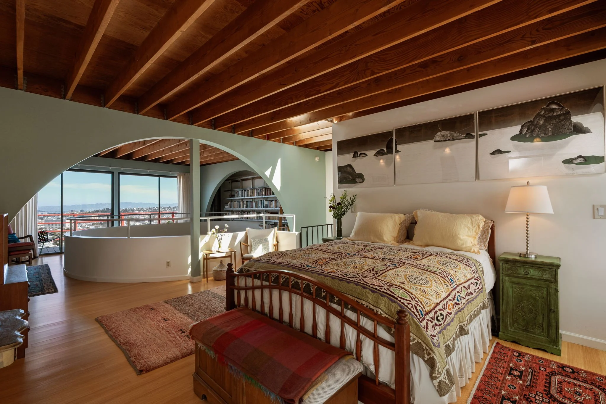 A bedroom with a wooden ceiling, a large bed with a patterned bedspread, green nightstand, lamp, and artwork of rocks and water on the wall. There is a window with a view of a cityscape and hills.