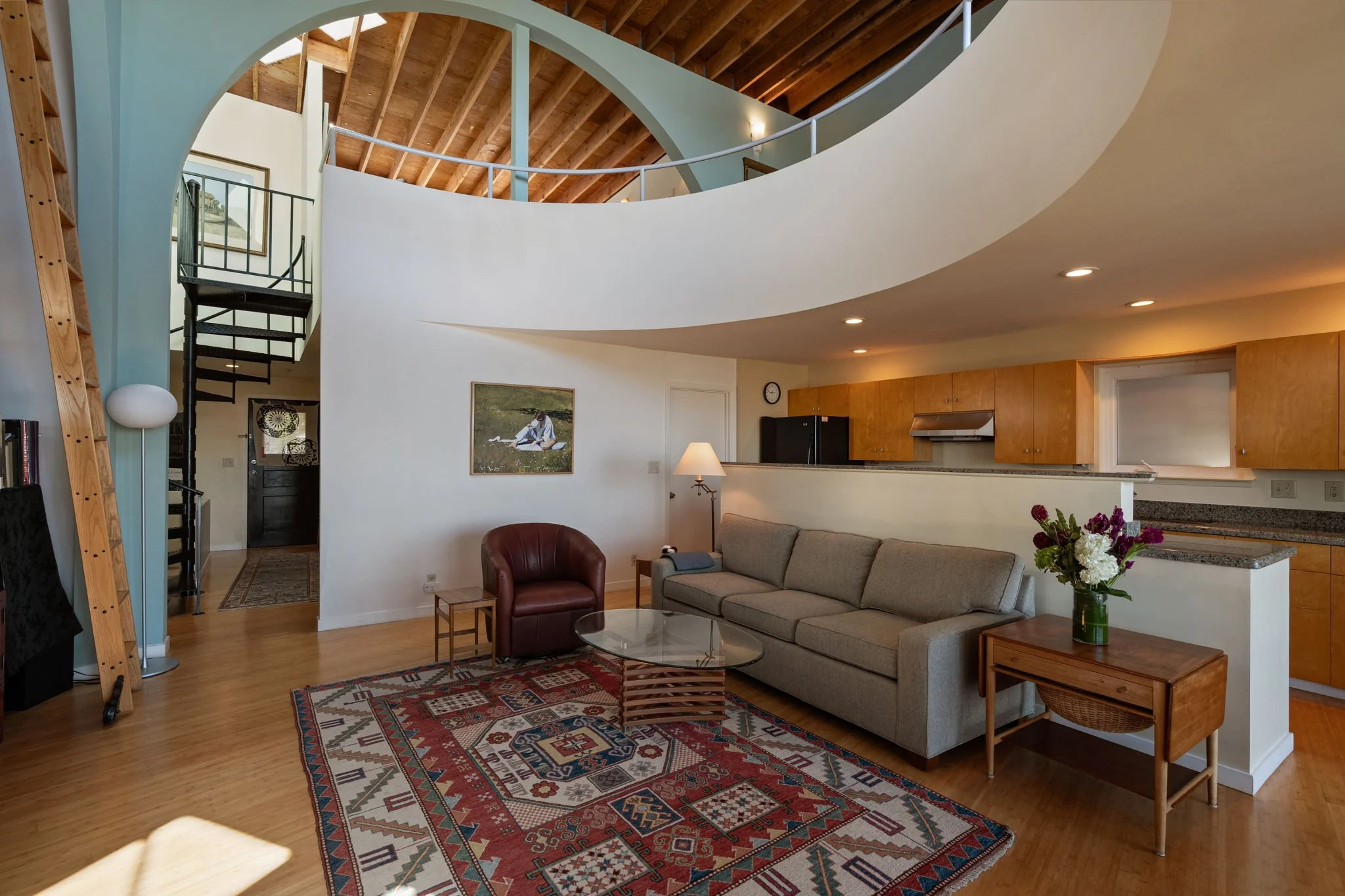 Living room with a gray sofa, glass coffee table, patterned rug, and a bouquet of flowers on a side table. Open kitchen with wooden cabinets and black refrigerator. Curved white wall with a staircase, wooden ceiling, and artwork on the wall.