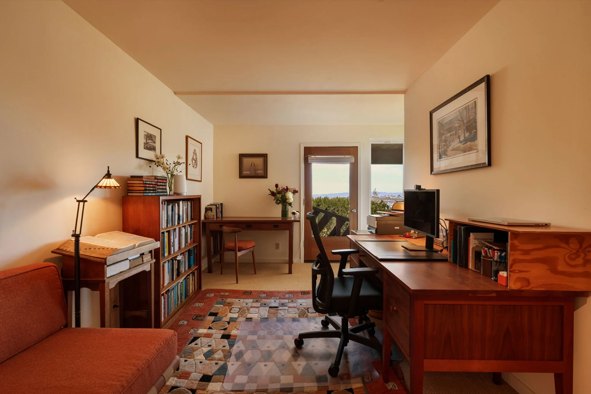 Living room with beige walls, wooden furniture, bookcases, framed pictures, a desk with a computer, a window with a view outside, and a patterned rug on the carpeted floor.