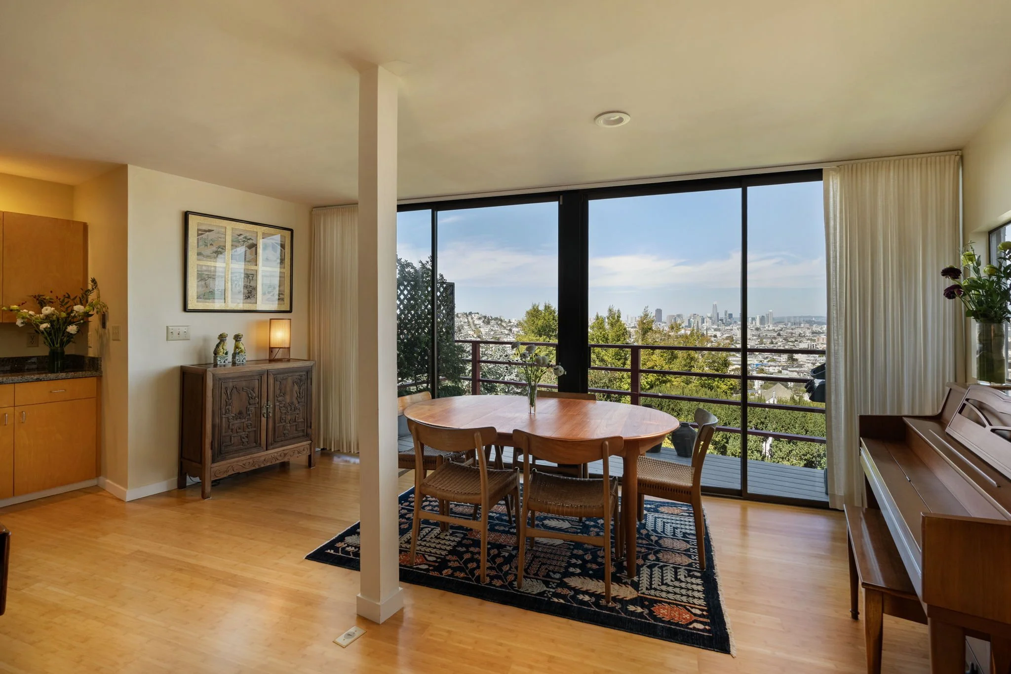 Living room with wooden floor, a dining table with chairs, a large window with a view of a city skyline and trees, a decorative cabinet, a framed picture on the wall, and various plants.
