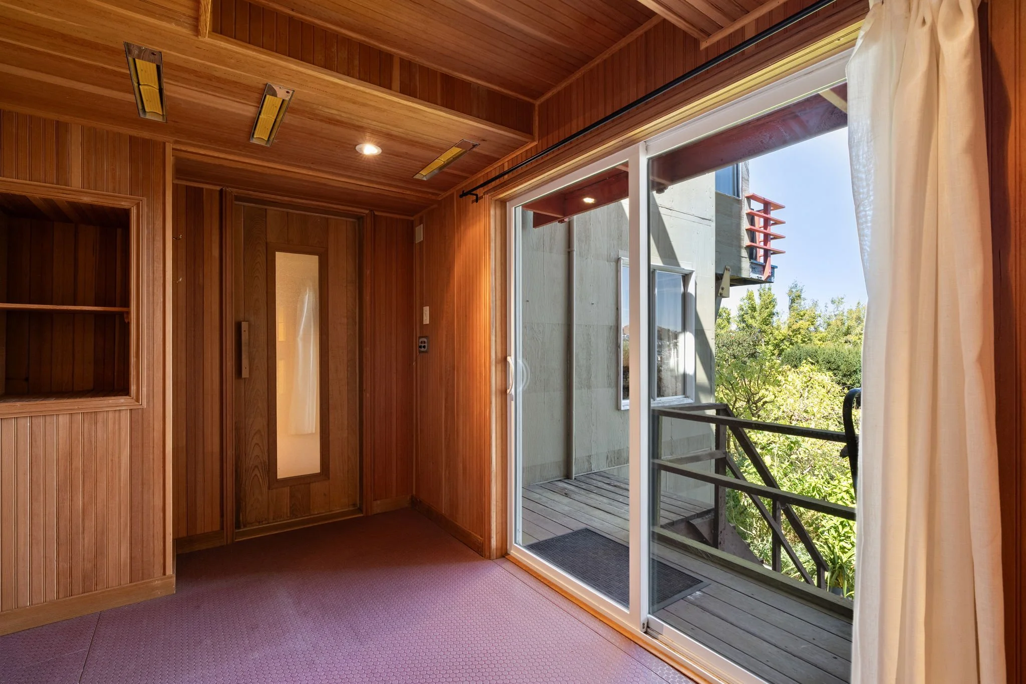 Interior view of a room with wood-paneled walls and ceiling, a sliding glass door leading to a balcony with outdoor stairs, and curtains on the right side.