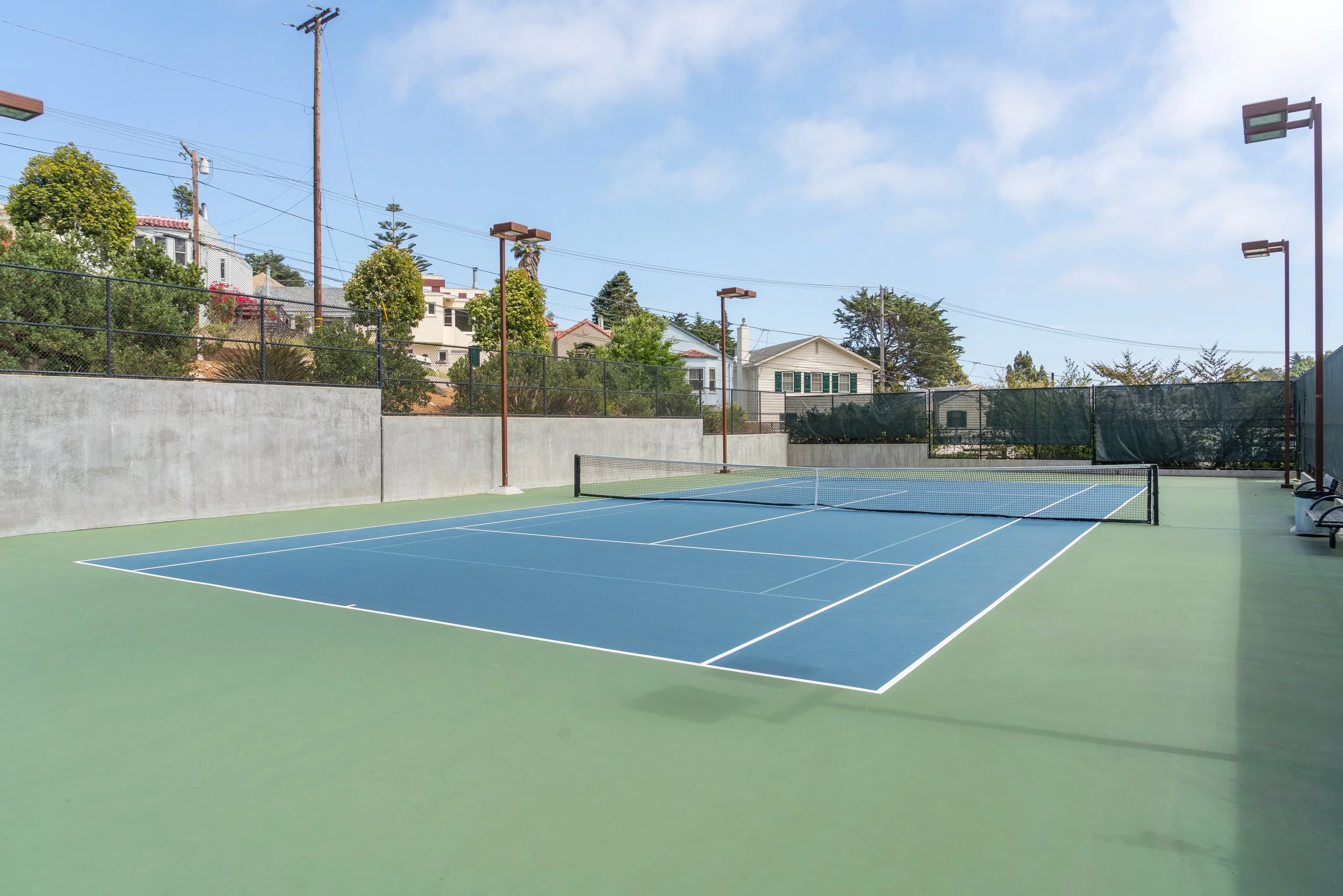 An outdoor tennis court with a blue playing surface, surrounded by a chain-link fence, under a clear sky with some clouds, and residential houses and trees in the background.