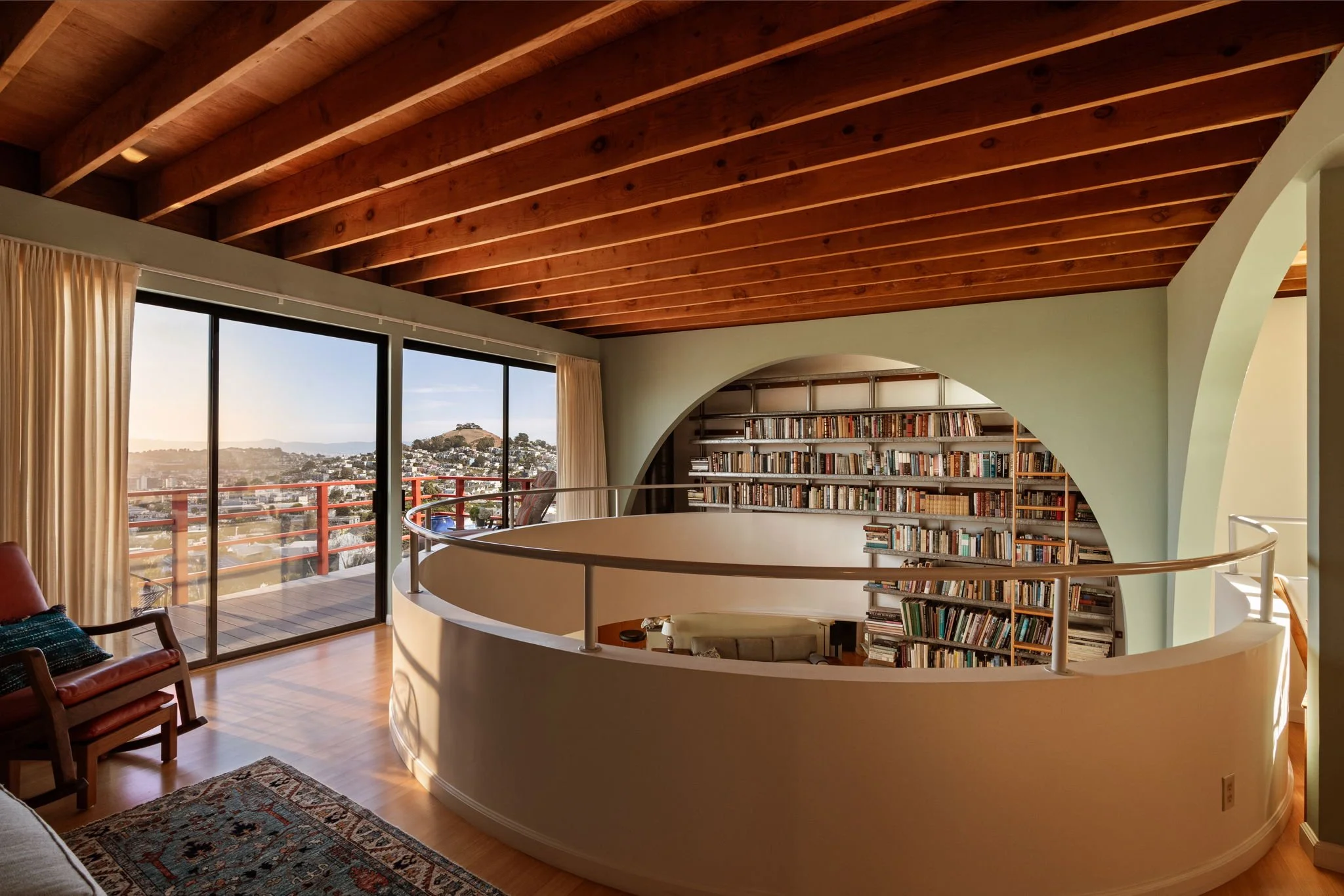 Interior view of a living room with a wooden beamed ceiling, large glass sliding doors opening to a balcony, and shelves filled with books. A curved wall surrounds a spiral staircase, and furniture includes chairs and a rug.