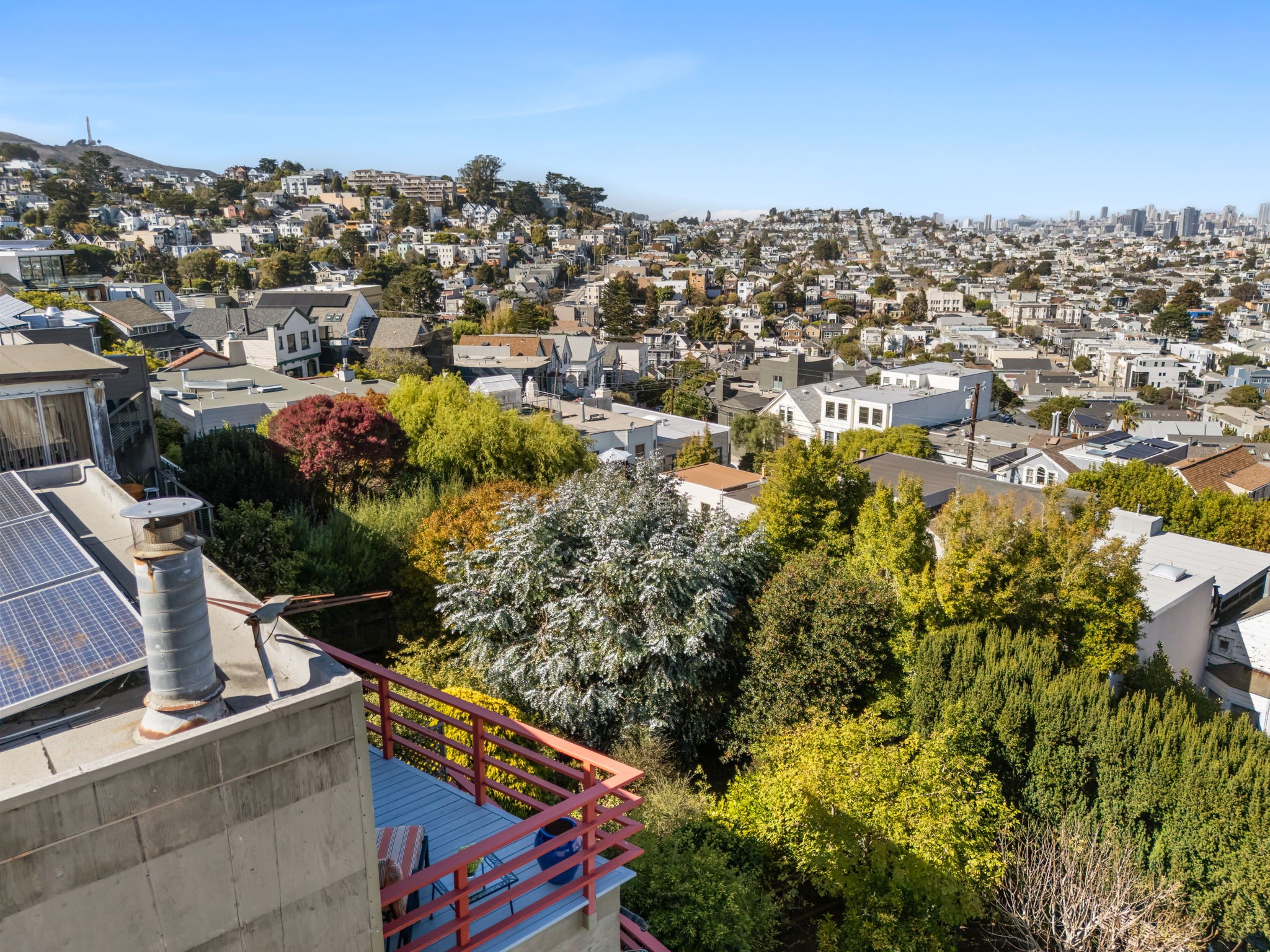 View from a hillside balcony overlooking a neighborhood with densely packed houses, trees, and a distant city skyline, under a clear blue sky.