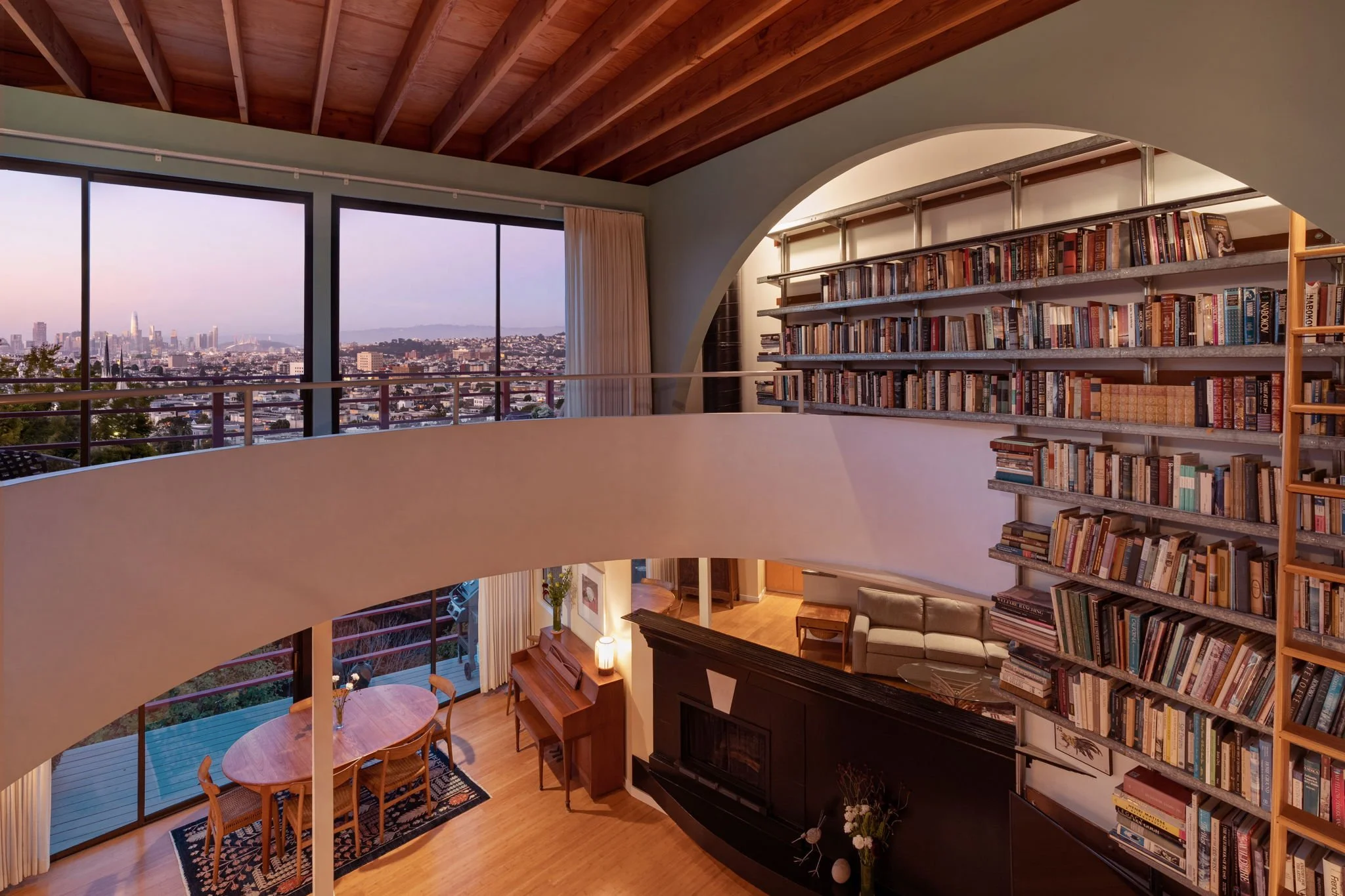 A spacious modern living room with large windows showing a cityscape view, a curved balcony railing, and a high ceiling with exposed wooden beams. There are bookshelves filled with books, a piano, and a dining table with chairs.