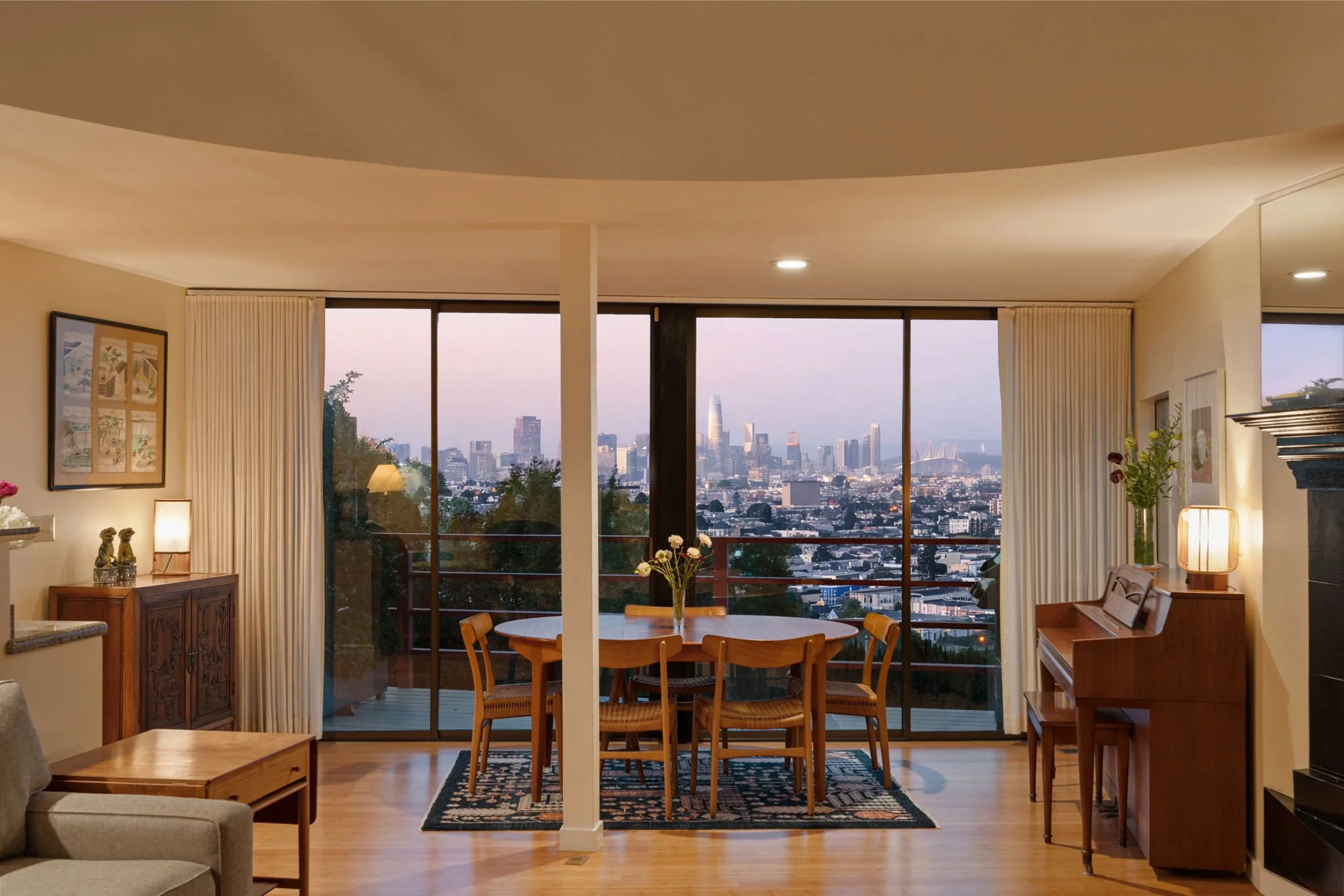 Living room with large windows showing city skyline at dusk, wooden dining table with chairs, potted plants, artwork, a piano, and various lamps.