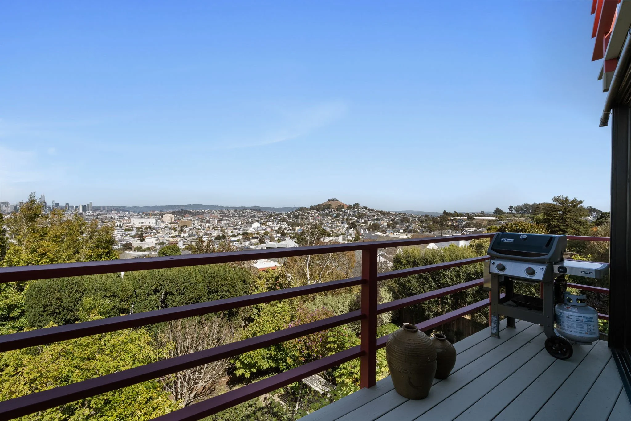 A balcony view overlooking a city skyline with a hill in the distance under a clear blue sky. There is a barbecue grill on the balcony, with large decorative pots and trees below.