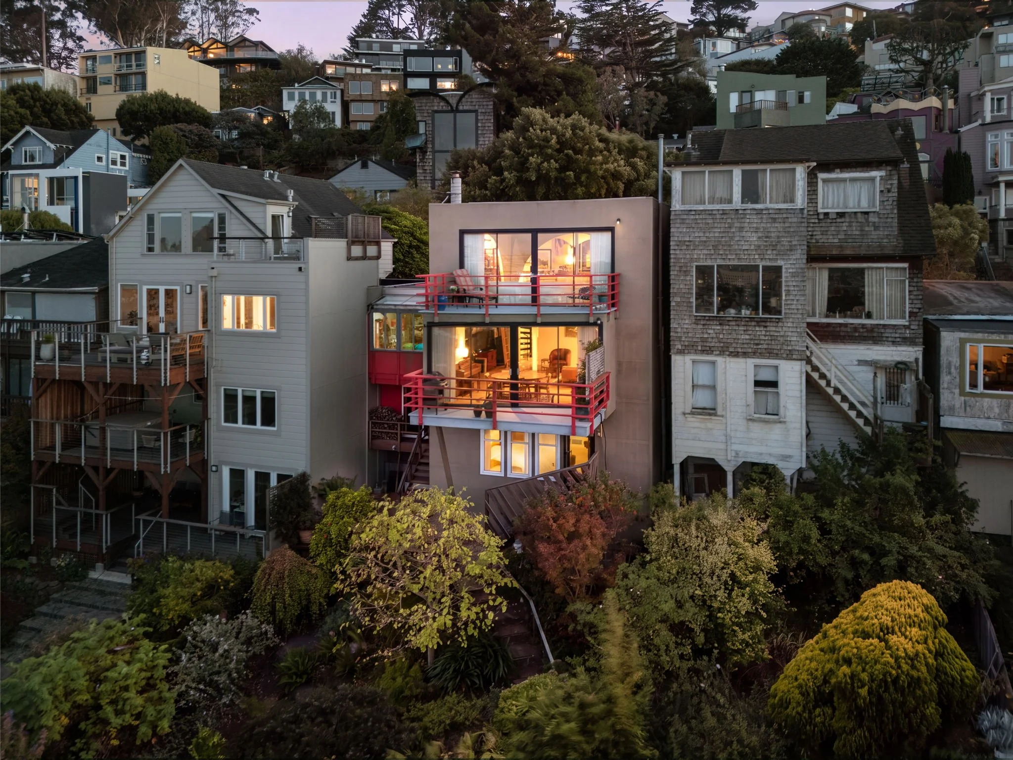 A hillside neighborhood with multiple modern houses, some with balconies, nestled among trees. The houses are illuminated with warm indoor lighting, and the sky indicates it is dusk.