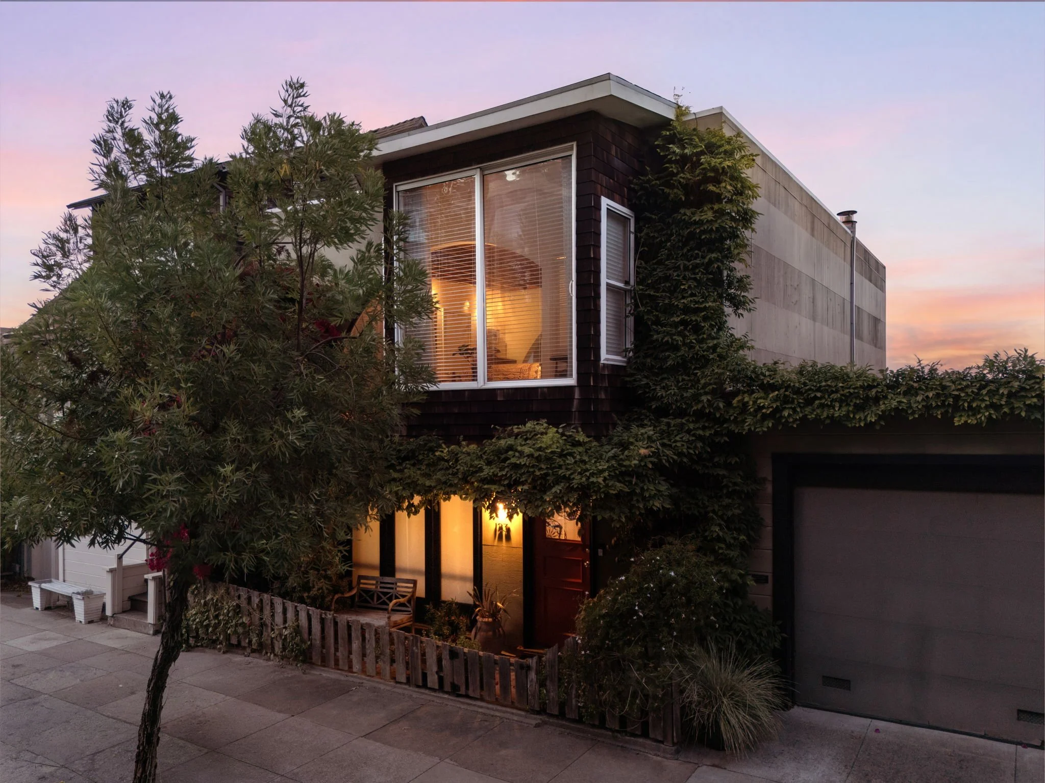 A modern two-story house with large windows and a mixture of dark and light exterior panels, surrounded by green trees at sunset.
