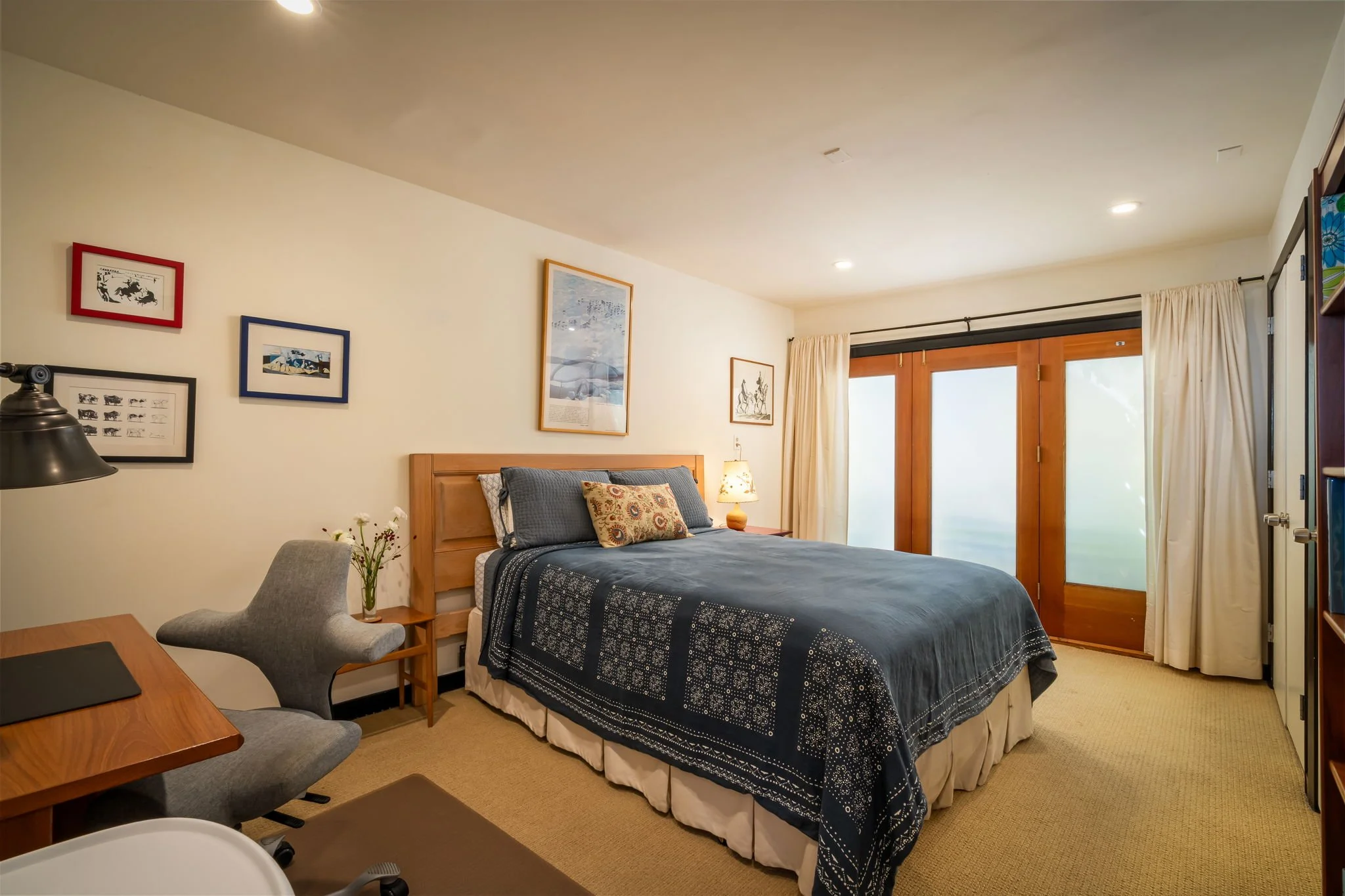 A cozy bedroom with a wooden bed frame, dark blue patterned bedding, and various decorative pillows. There is a nightstand with a lit lamp and artwork hanging on the walls. A large sliding glass door leads to a balcony or patio, framed by white curta