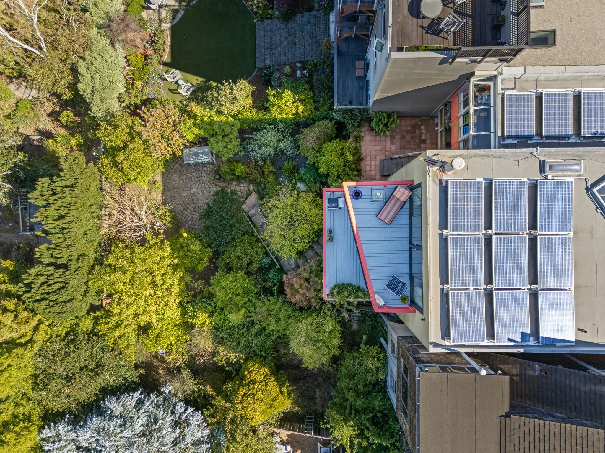 An aerial view of a backyard with lush greenery, a stone pathway, and patio furniture adjacent to a house with solar panels on the roof.