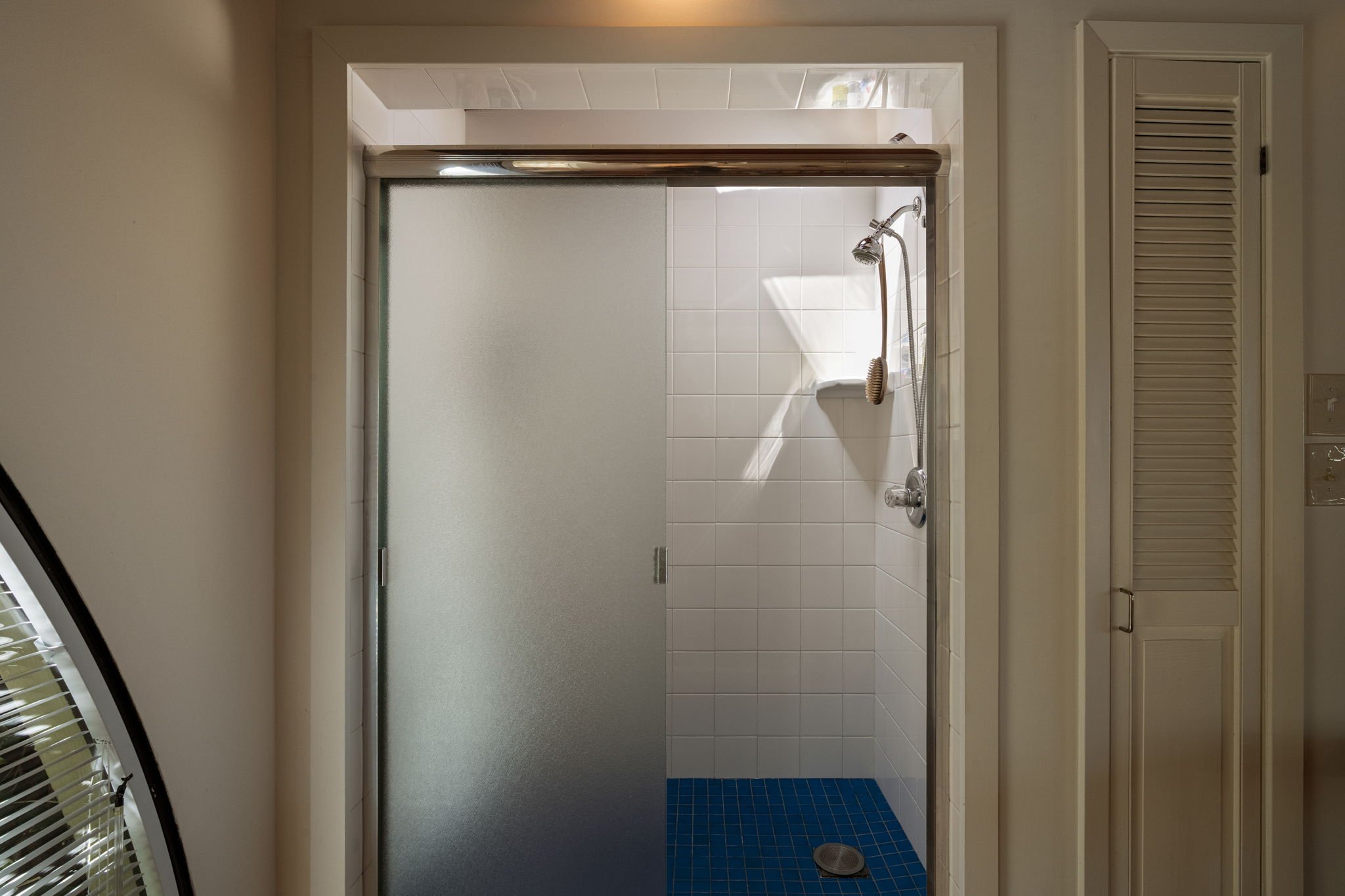 View of a small shower with white tiles on the wall and a blue tiled floor with a drain, and a silver showerhead with a flexible hose, seen through a glass door.