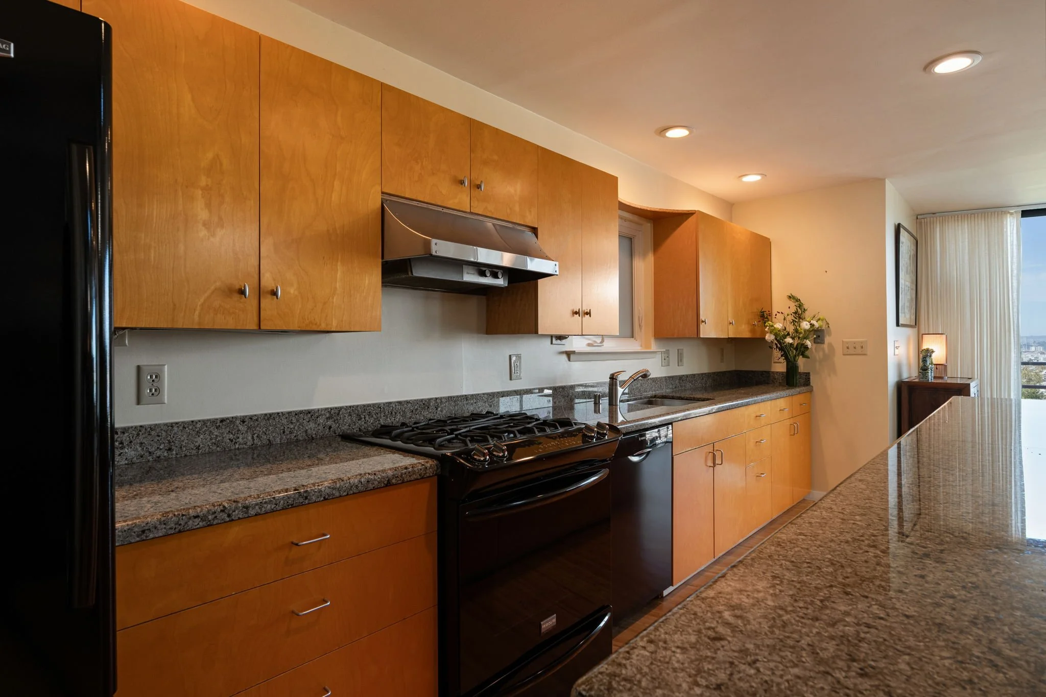 Kitchen with wooden cabinets, granite countertop, black stove, stainless steel range hood, and a black refrigerator. There is a window with curtains and a small table with a flower vase in the background.