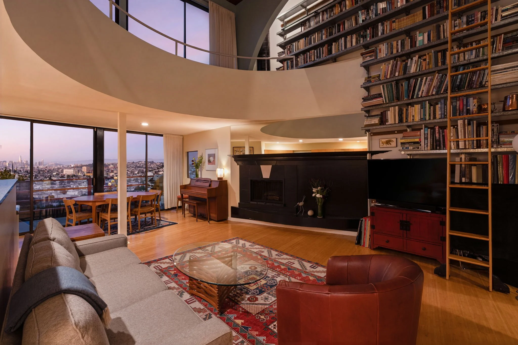 Living room with large windows, city skyline view, a gray sofa, glass coffee table, red leather armchair, wooden dining table and chairs, a bookshelf filled with books, a piano, and indoor plants.