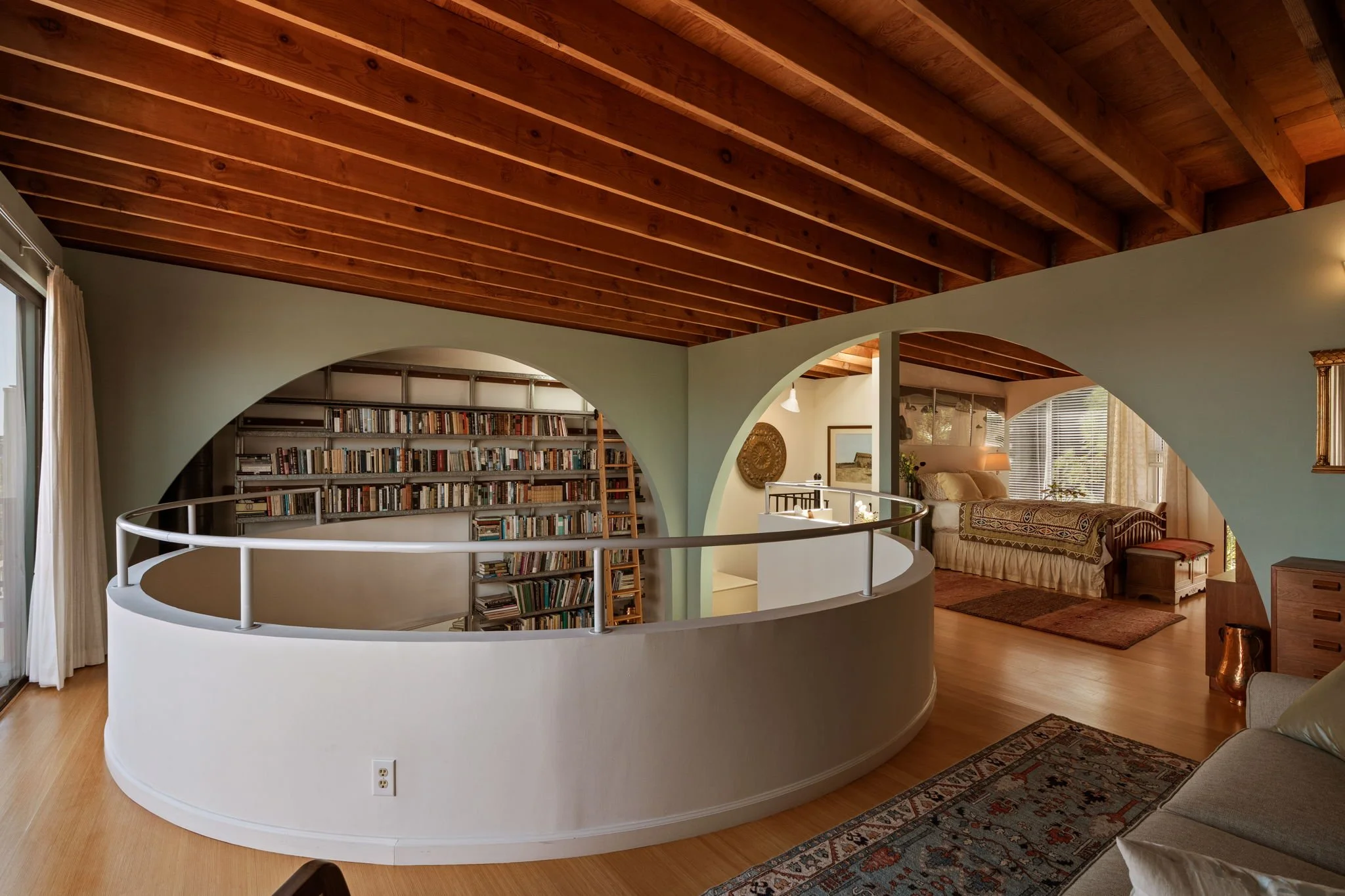 Open living room with wooden ceiling, curved white railing, bookshelves, sitting area with sofa, and a large window with blinds.