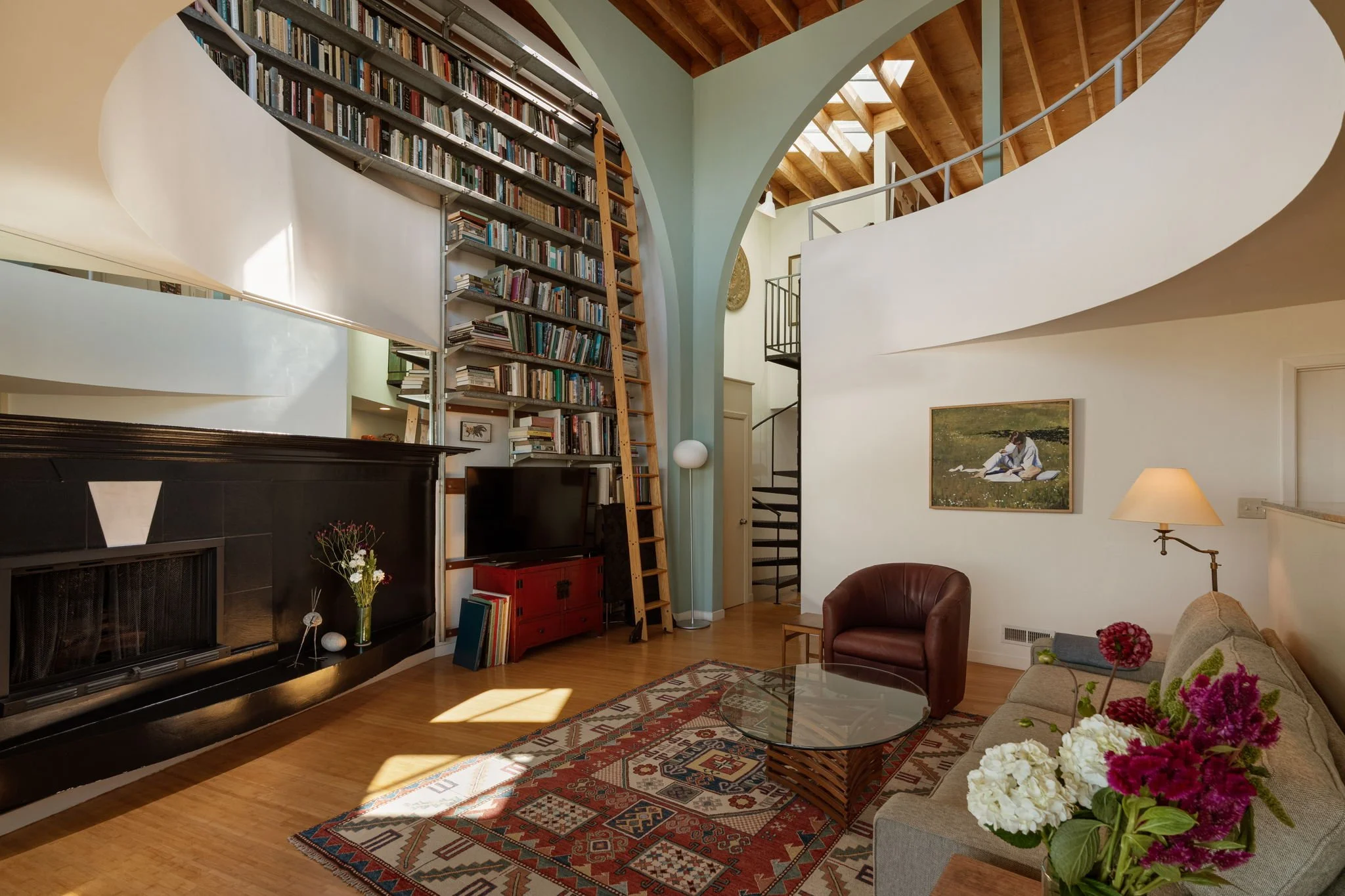A spacious living room with a high ceiling featuring wooden beams and an arched wall with a large bookshelf reaching up to the ceiling. The room has a black fireplace, a red cabinet, a leather armchair, a corner with a lamp, and a sofa with flowers o