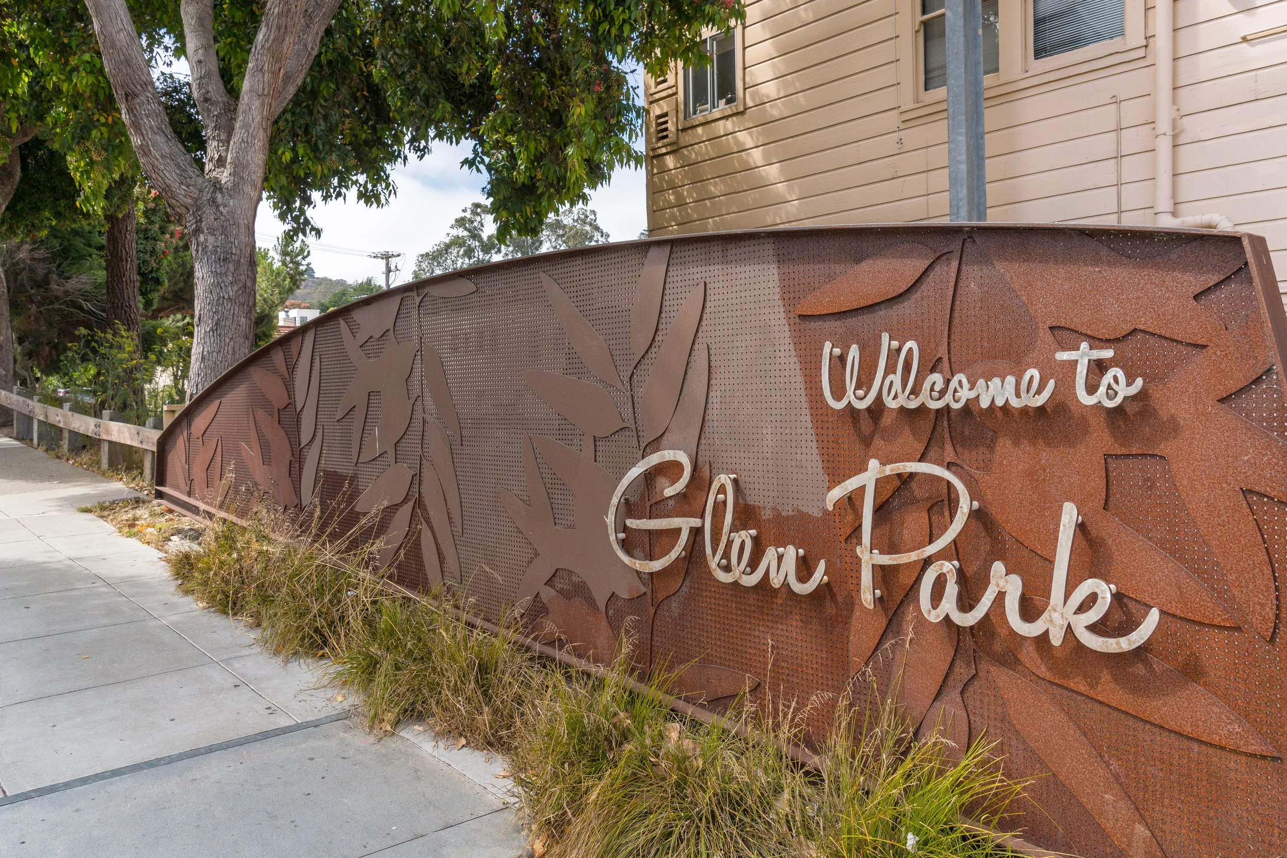 A decorative sign with the words "Welcome to" and other text that is partially obscured, mounted on a rust-colored metal panel with leaf patterns. There are trees and a beige building with windows in the background.