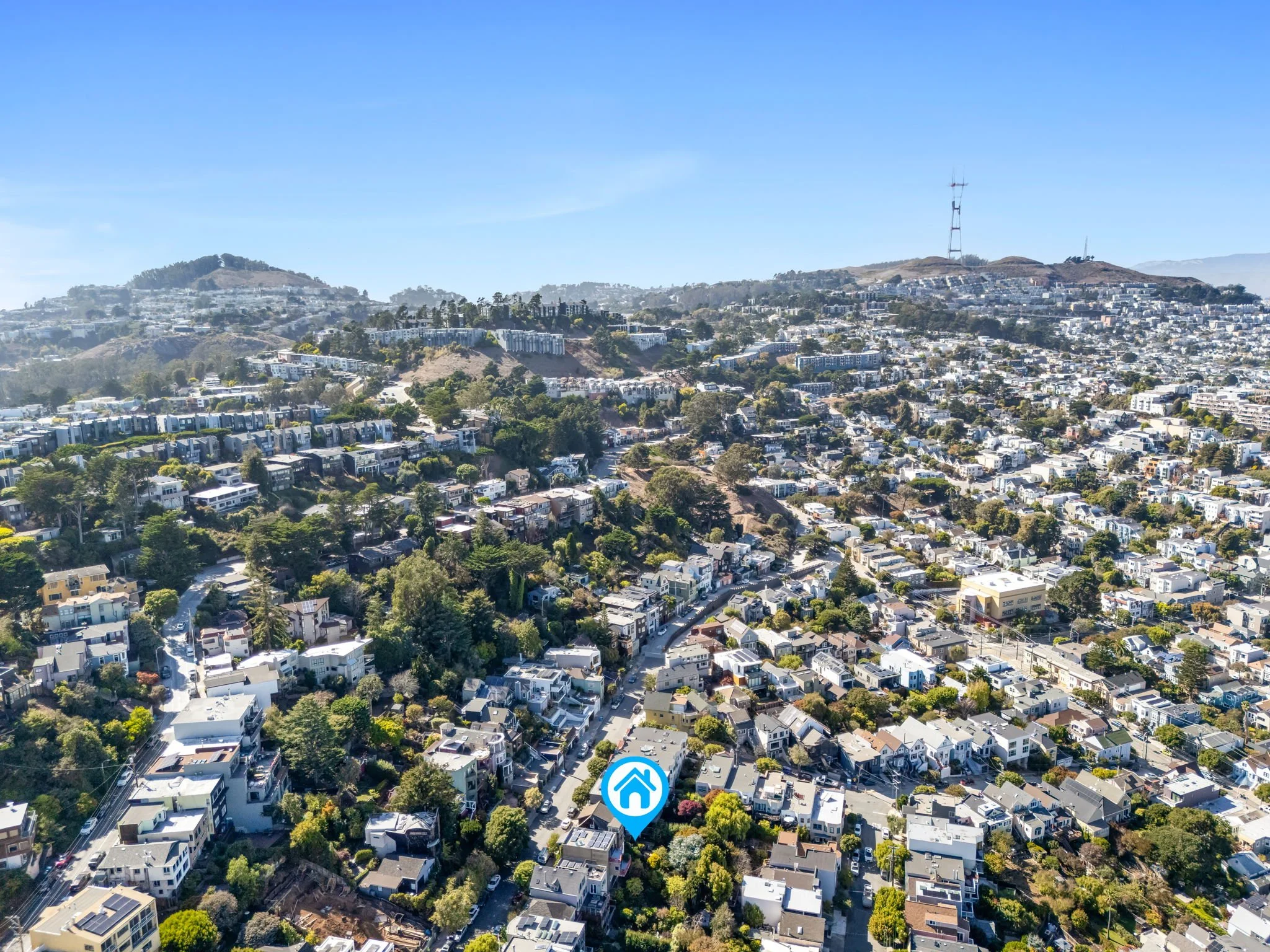 Aerial view of a densely populated residential neighborhood with houses, trees, and hills in the background under a clear blue sky.