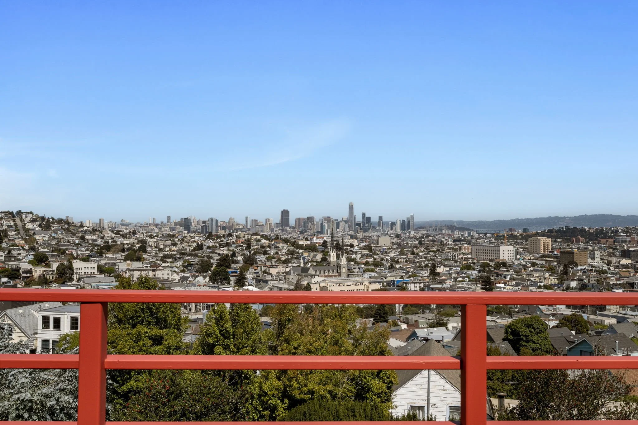 View of San Francisco city skyline from a balcony with red railing, featuring numerous buildings, including the Transamerica Pyramid, under a clear blue sky.