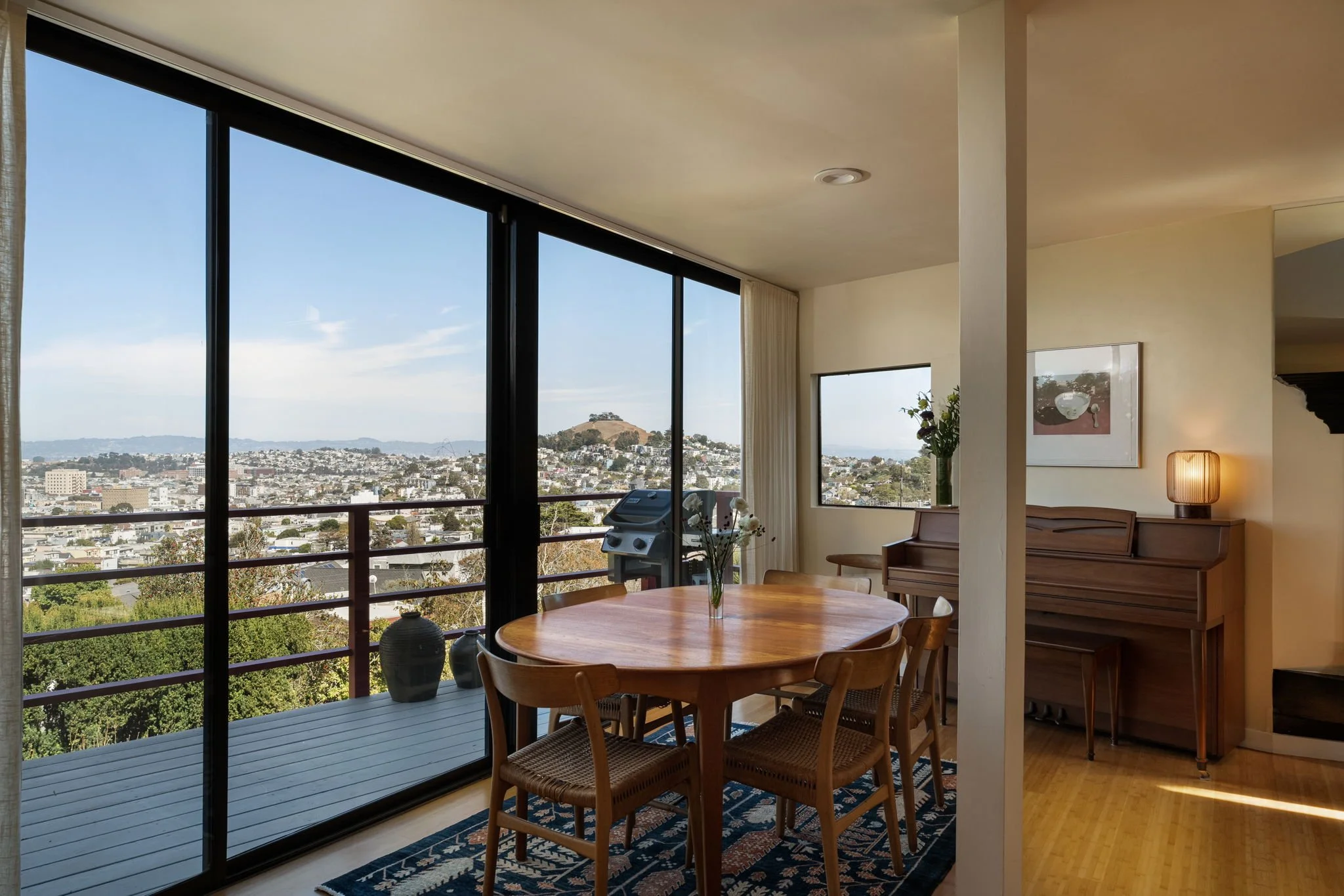 Interior of a living room with large glass sliding doors showing a city view with hills in the background, a round wooden dining table, chairs, a cushioned bench, a piano, a floor lamp, and decorative vases on the table.