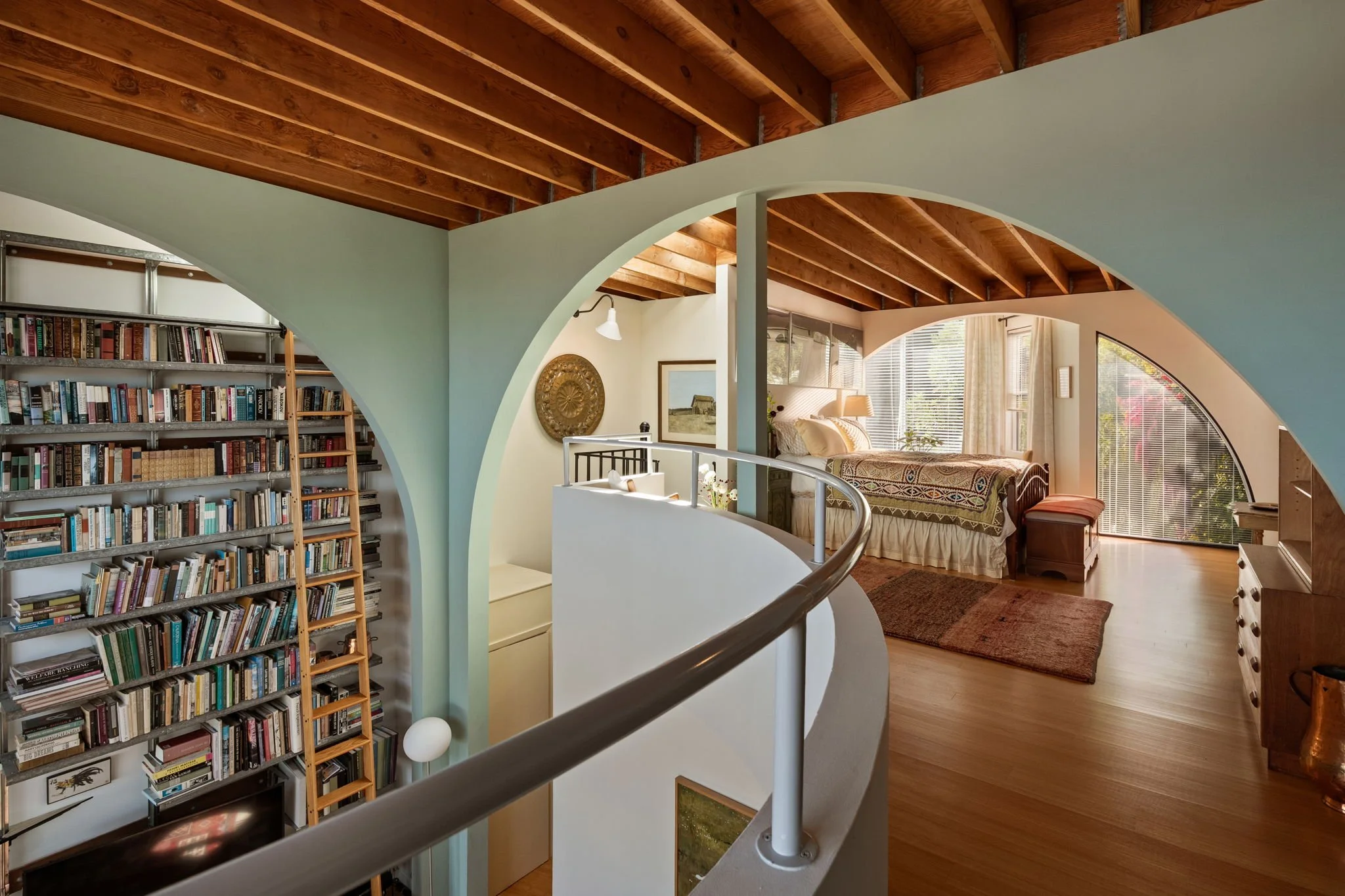 Interior view of a home with a curved staircase, a large bookshelf on the left, and a bedroom with a bed, windows, and curtains in the background. The ceiling has exposed wooden beams.