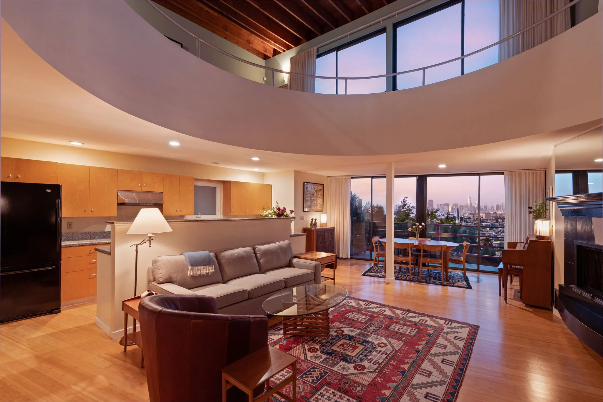 Living room and kitchen area with large windows, city skyline view, wooden ceiling, and modern furniture including a gray sofa, coffee table, dining table with chairs, and open floor plan