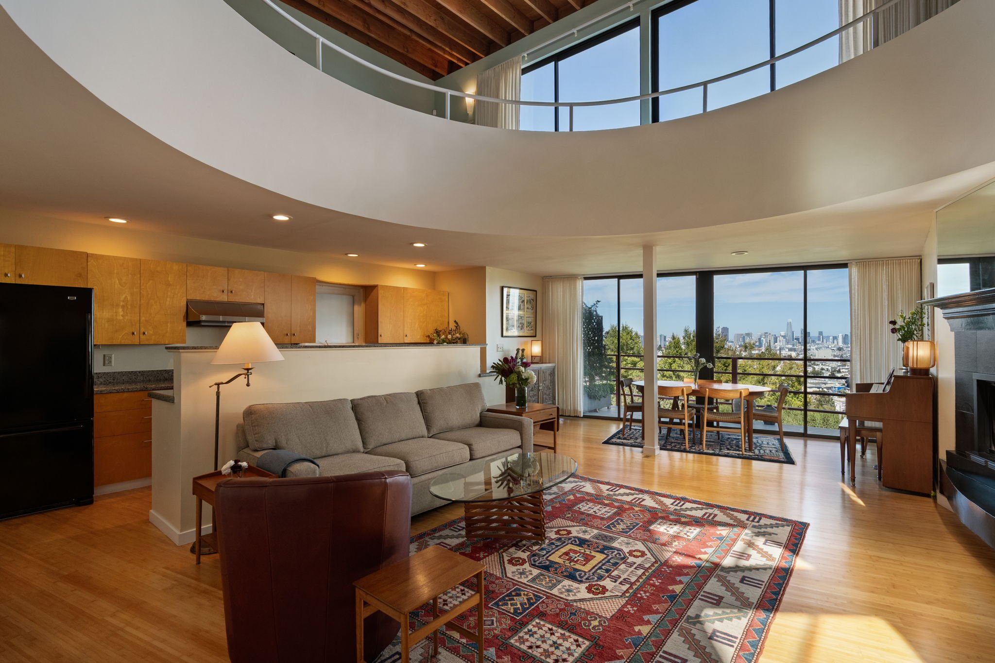 Interior of a modern apartment living room with large windows showing a city view, a beige sectional sofa, a glass coffee table, wooden kitchen cabinets, a patterned rug, and a dining area with a round table and four chairs.